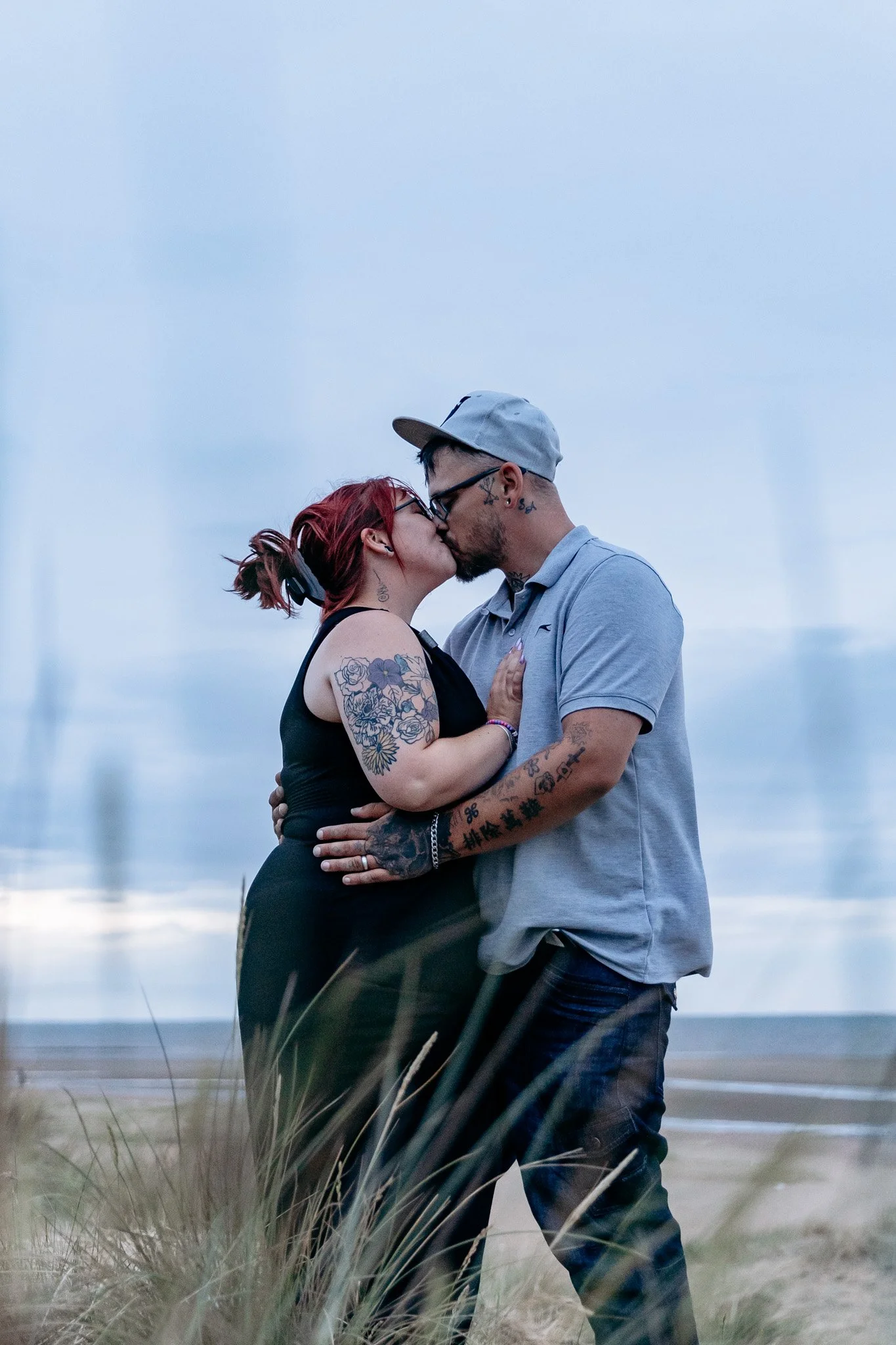 A couple sharing a kiss outdoors during overcast weather, with grassy dunes in the foreground.