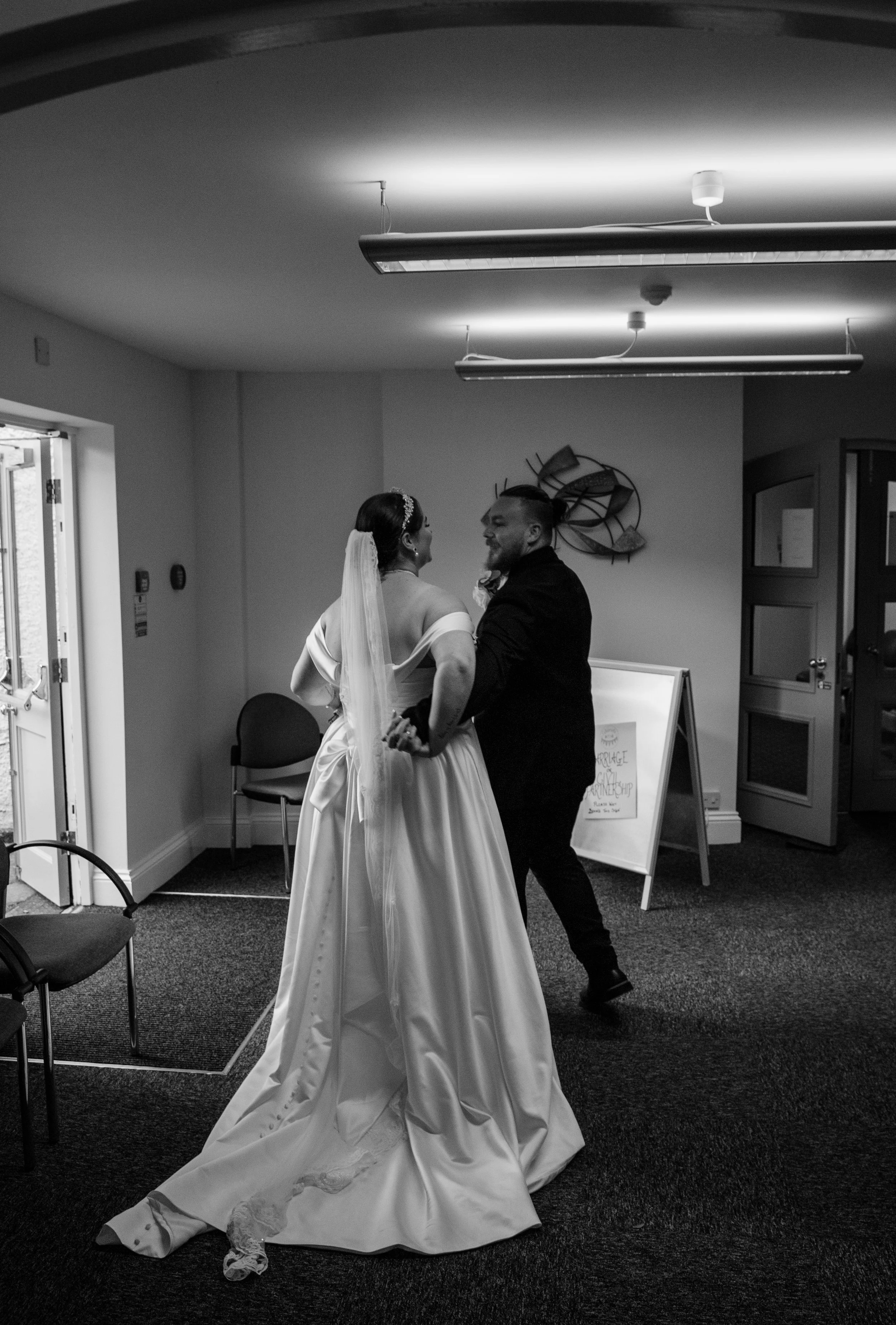 A bride in a wedding dress and a groom in a suit dancing together indoors, smiling at each other.