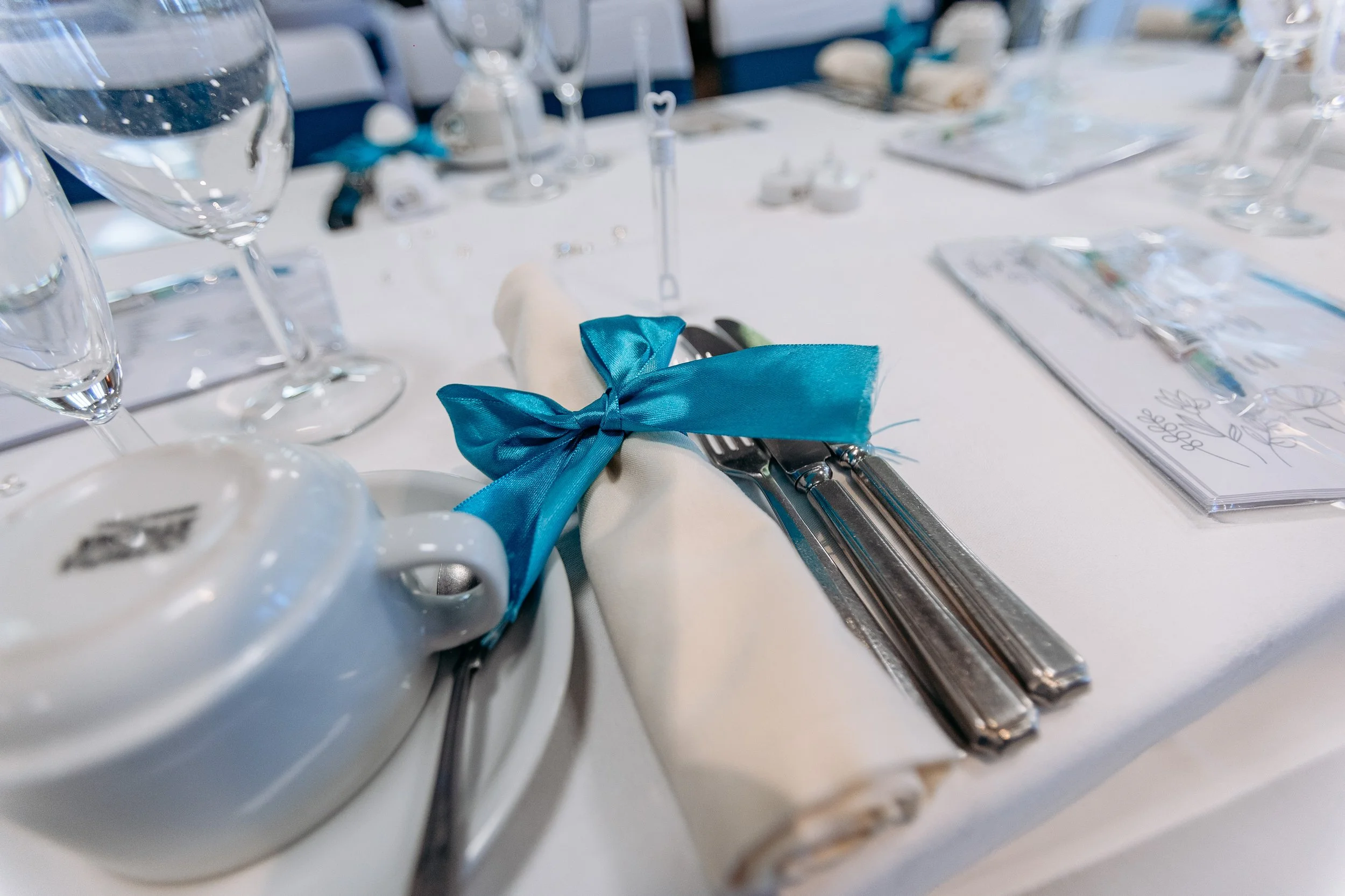 Wedding table setting with a white napkin tied with a blue ribbon, silverware, glassware, and a menu on a white tablecloth.