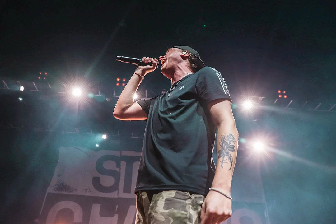 A person singing into a microphone on stage with bright lights in the background.