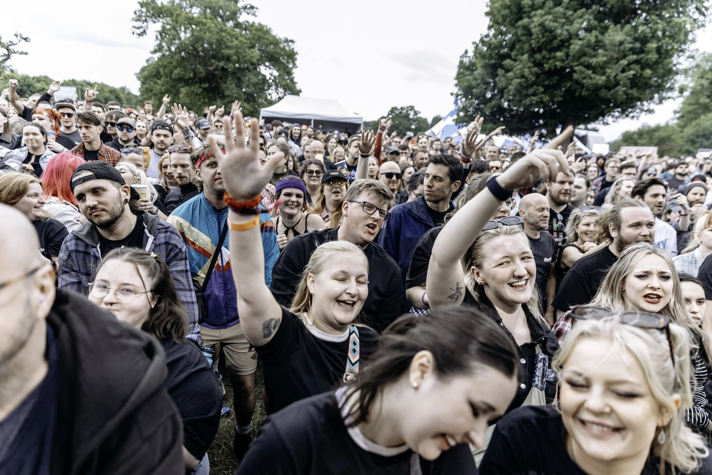 Crowd of people at an outdoor concert or festival, smiling and enjoying themselves, with some raising their hands.