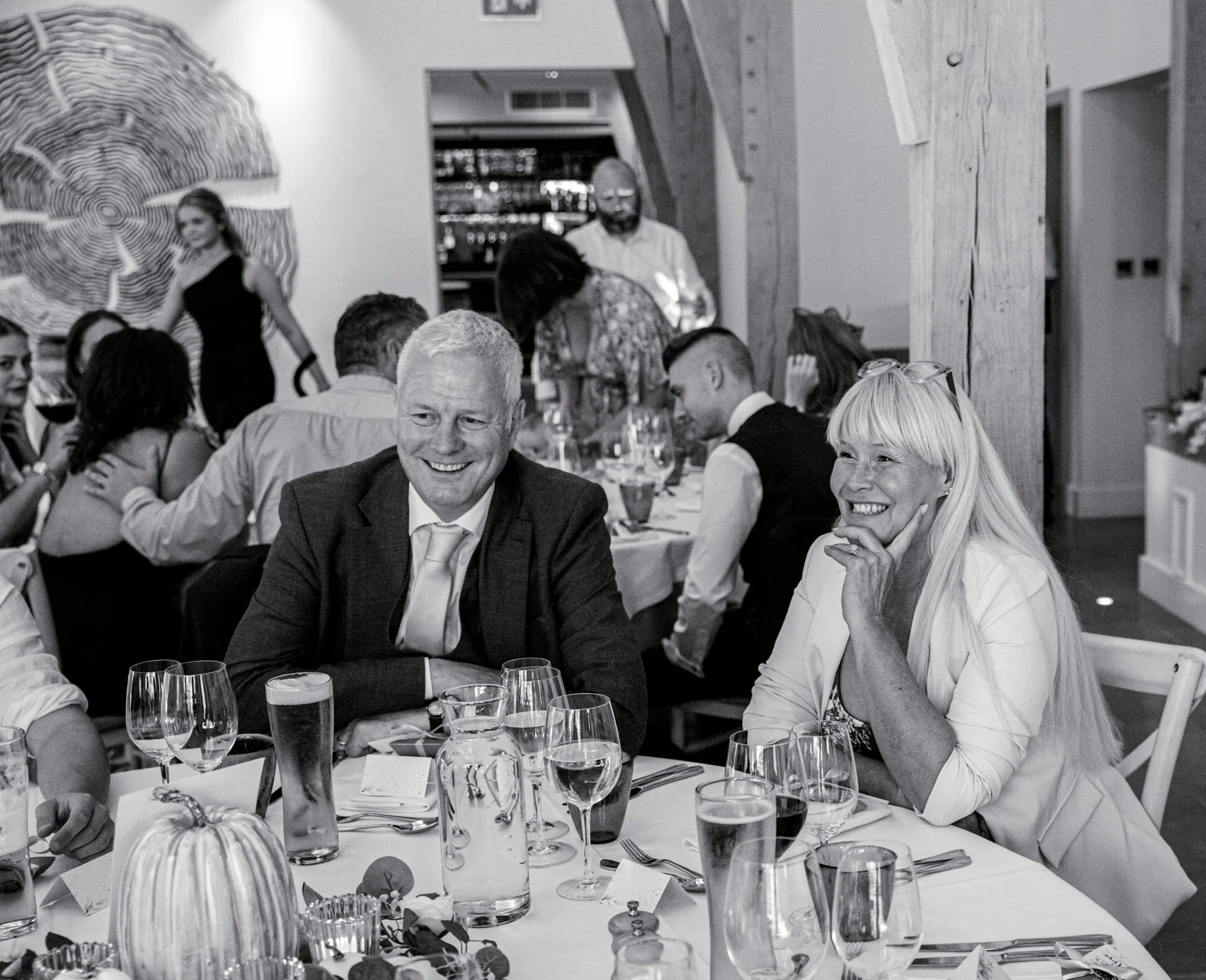Black and white photo of people seated at a dining table during a social event, featuring a man and woman smiling, with glasses and decorative items on the table.