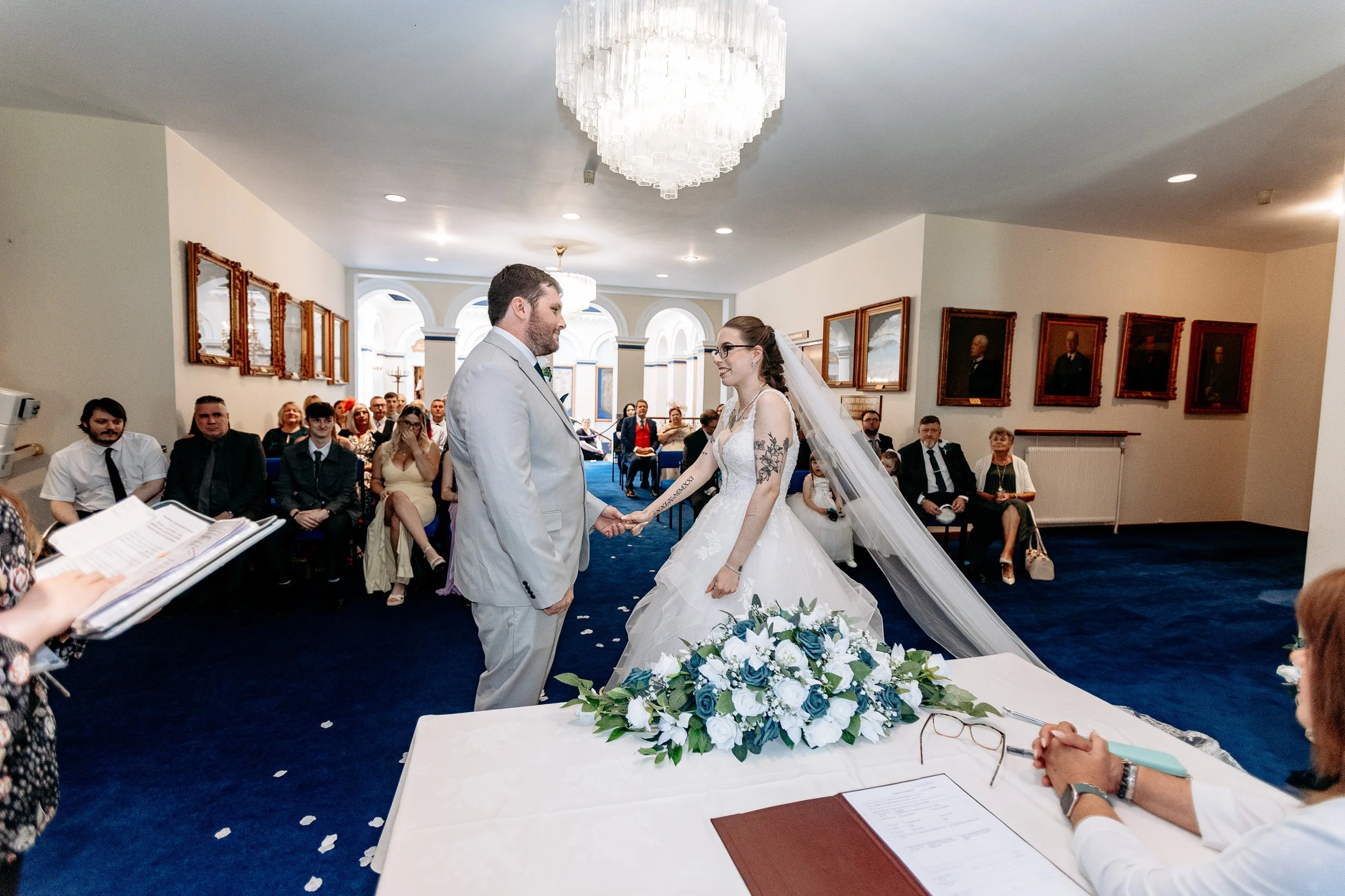 A bride and groom holding hands during their wedding ceremony in a decorated indoor venue with seated guests watching.