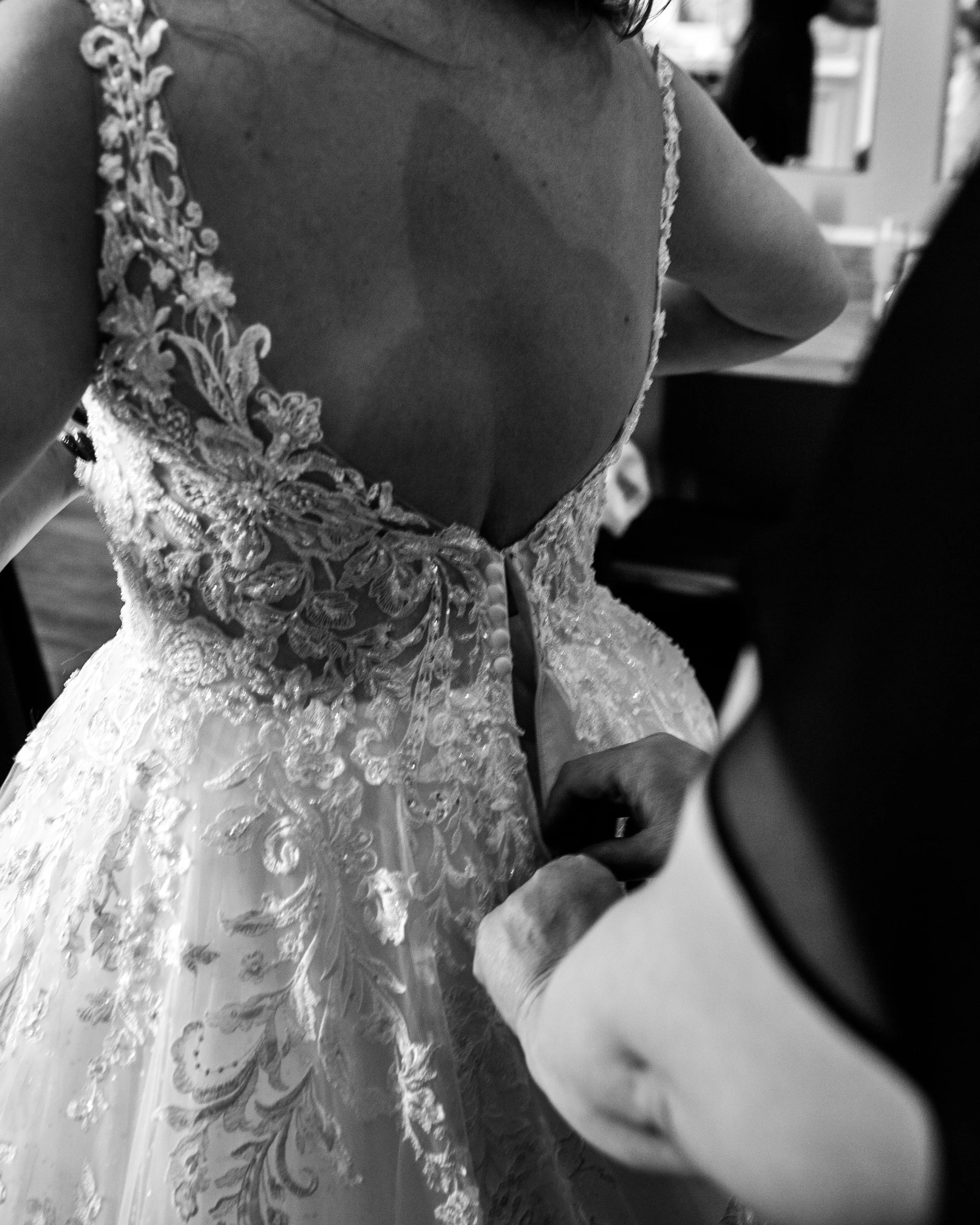 A woman in a wedding dress is being fastened at the back, with intricate lace detailing and buttons, as someone helps her prepare for her wedding.