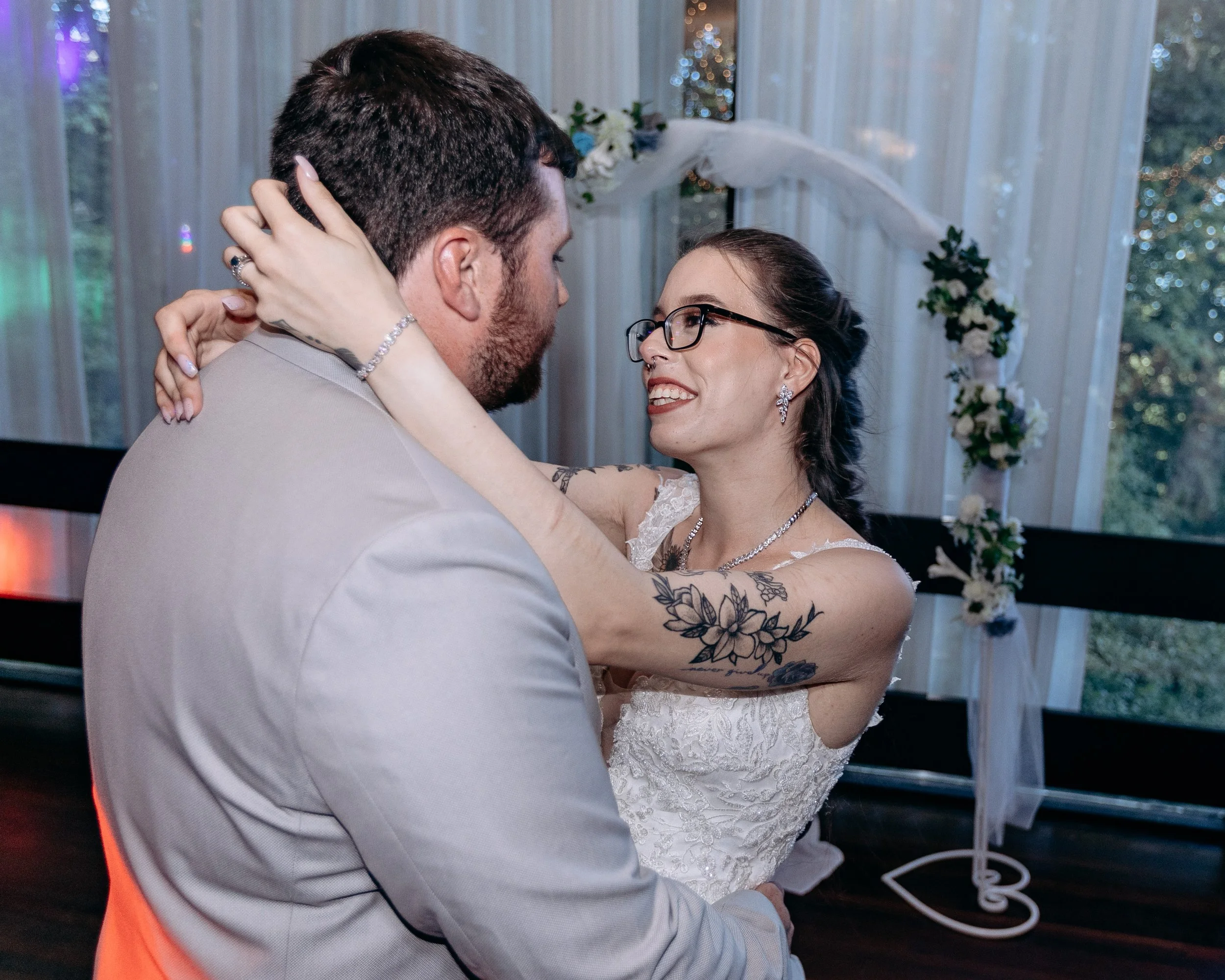 A bride and groom dancing closely at their wedding reception, smiling at each other, in front of a decorated floral arch and large windows with curtains.