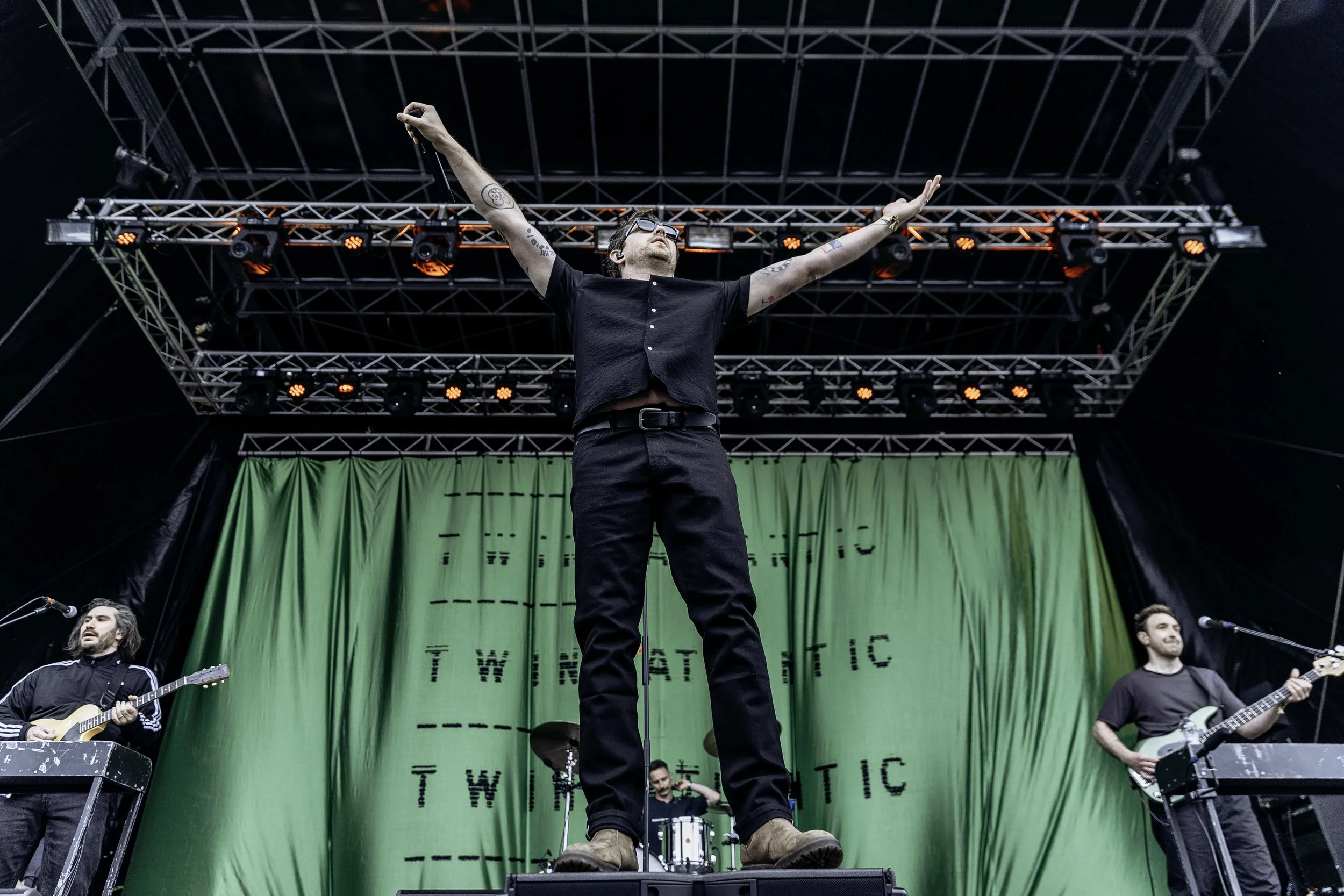 A live music band performing on a stage with a green backdrop, with the lead singer standing at the front with arms raised. The band members are playing guitars, and there is a drummer in the background. The stage has multiple lights and scaffolding 