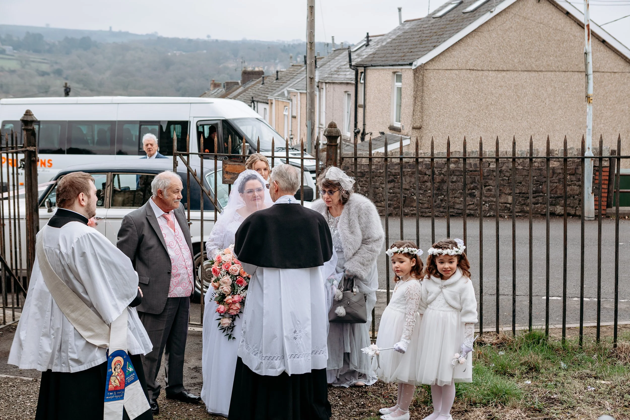 Group of people gathered outside near a fence, including two young girls in white dresses with flower crowns, a bride in a white wedding gown with veil, and several adults, some dressed as clergy or in formal attire, during a wedding ceremony.