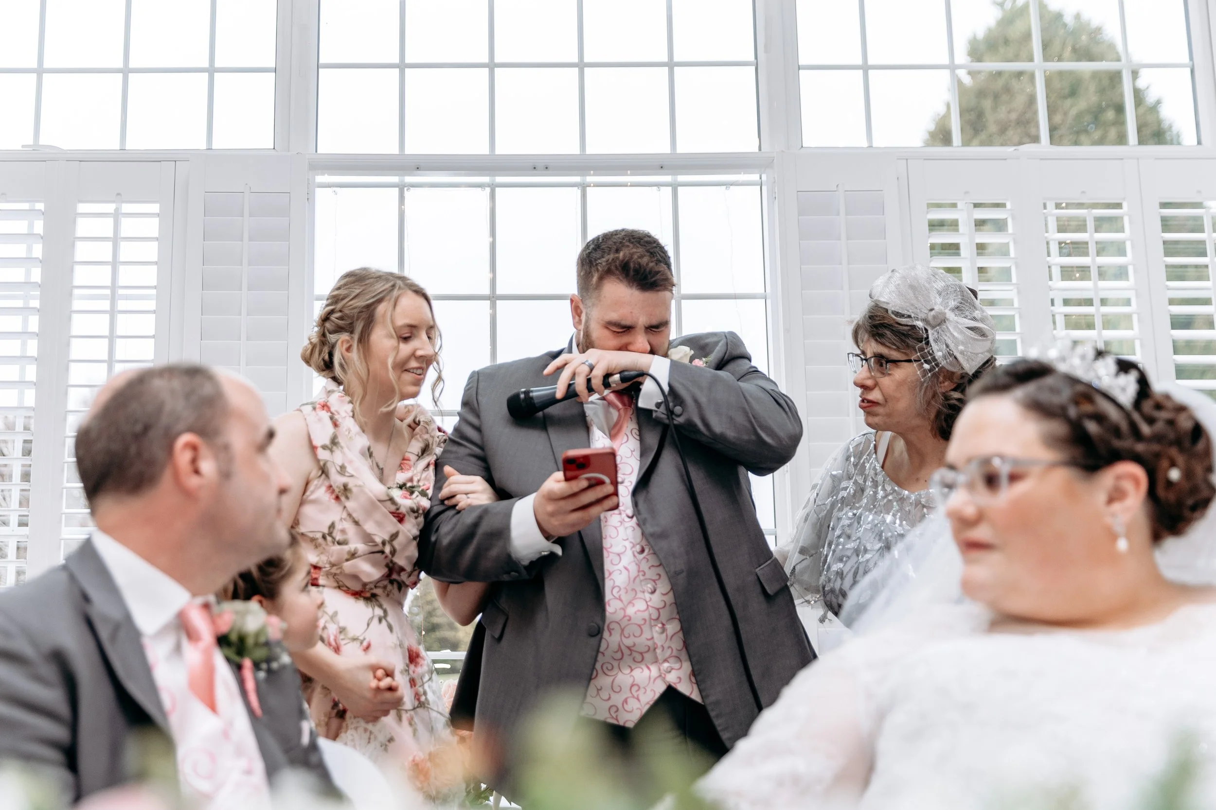 A man in a suit appears to be crying or emotional during a wedding ceremony, holding a microphone and a phone, surrounded by women and a girl, with a large window with white shutters in the background.