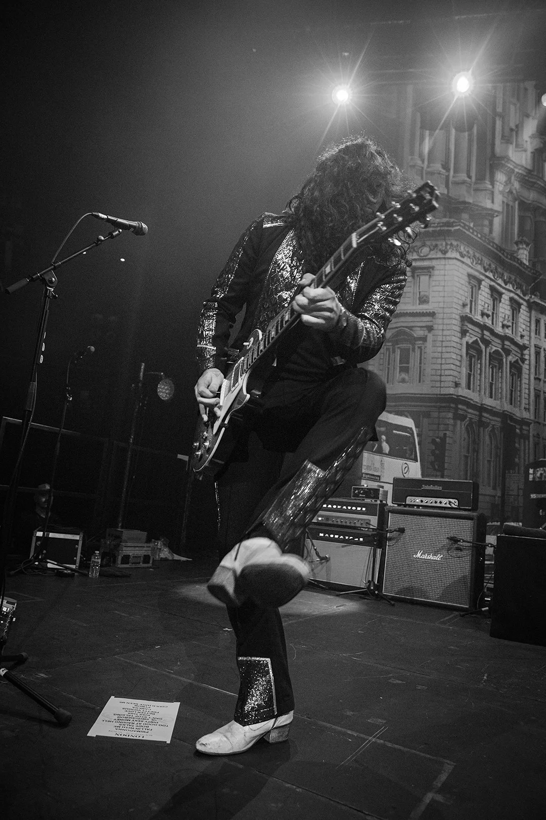 Black and white photo of a guitarist performing energetically on stage, wearing a glittery suit with a bent knee, surrounded by amplifiers and a microphone stand, with bright stage lights and a building backdrop.