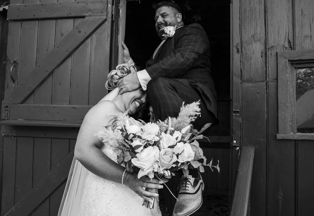 A bride and groom laughing and hugging at their wedding, with the groom sitting on a wooden barn door frame and the bride holding a bouquet of flowers, in a black and white photograph.