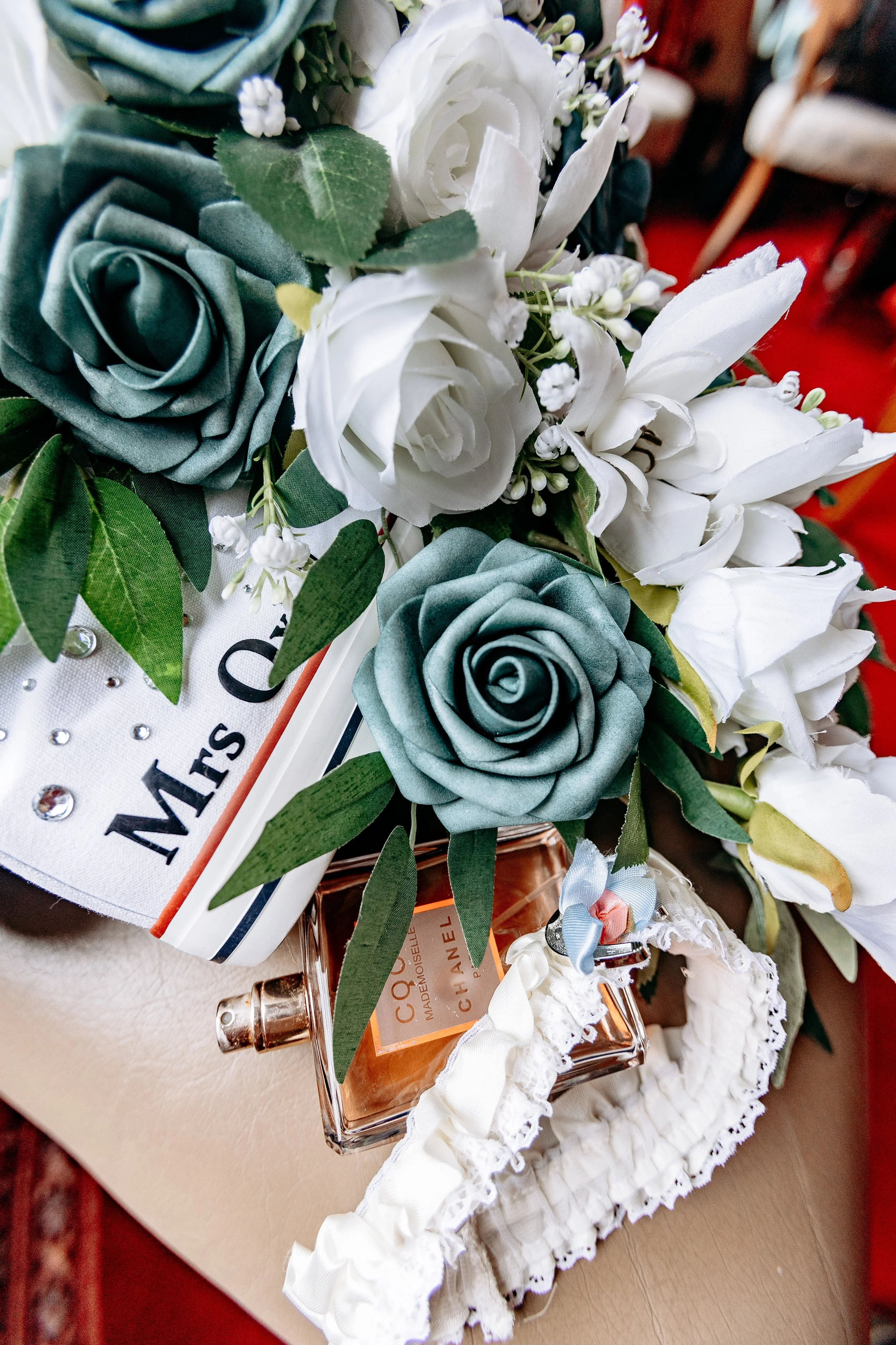A bouquet of white and blue artificial roses, white lilies, and greenery with a decorative ribbon, alongside a bottle of Chanel Coco Mademoiselle perfume, and a white lace garter with a blue bow, arranged on a surface with a sign reading 'Mrs.'