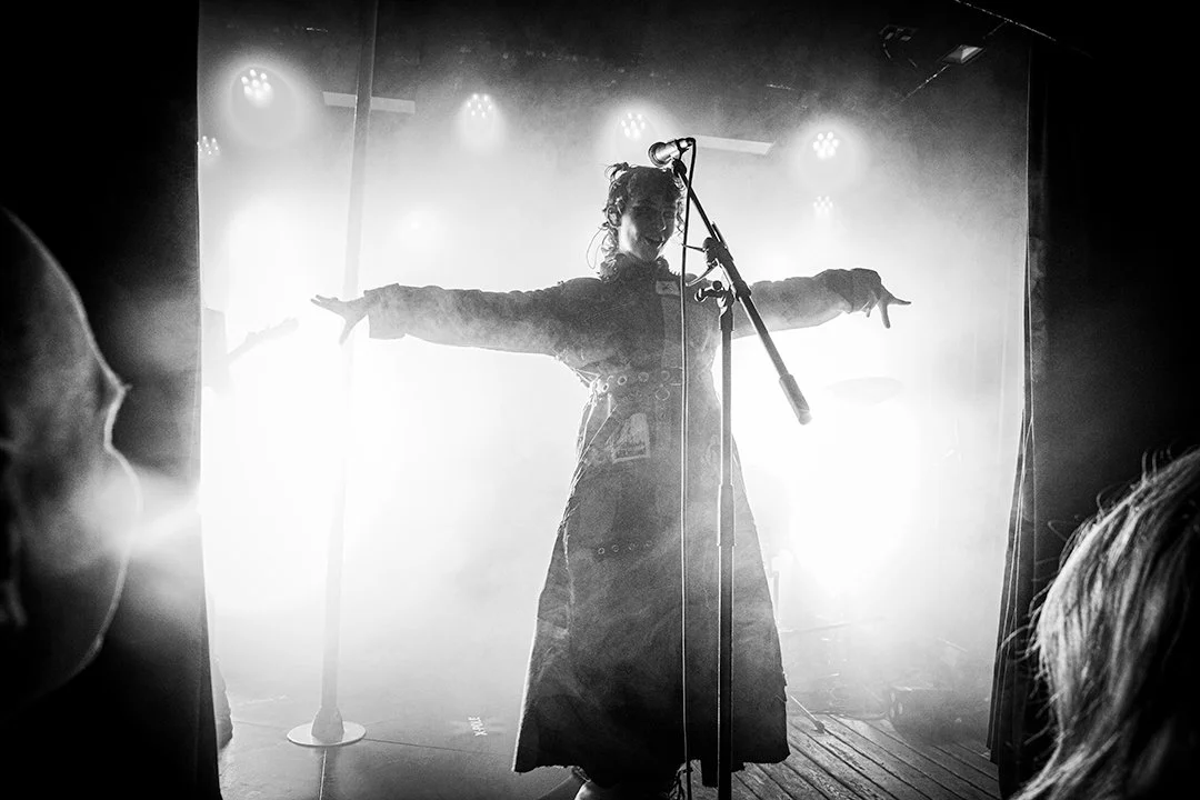 A woman standing on stage with arms outstretched, microphone in front, backlit by bright lights, surrounded by audience members in a concert setting.
