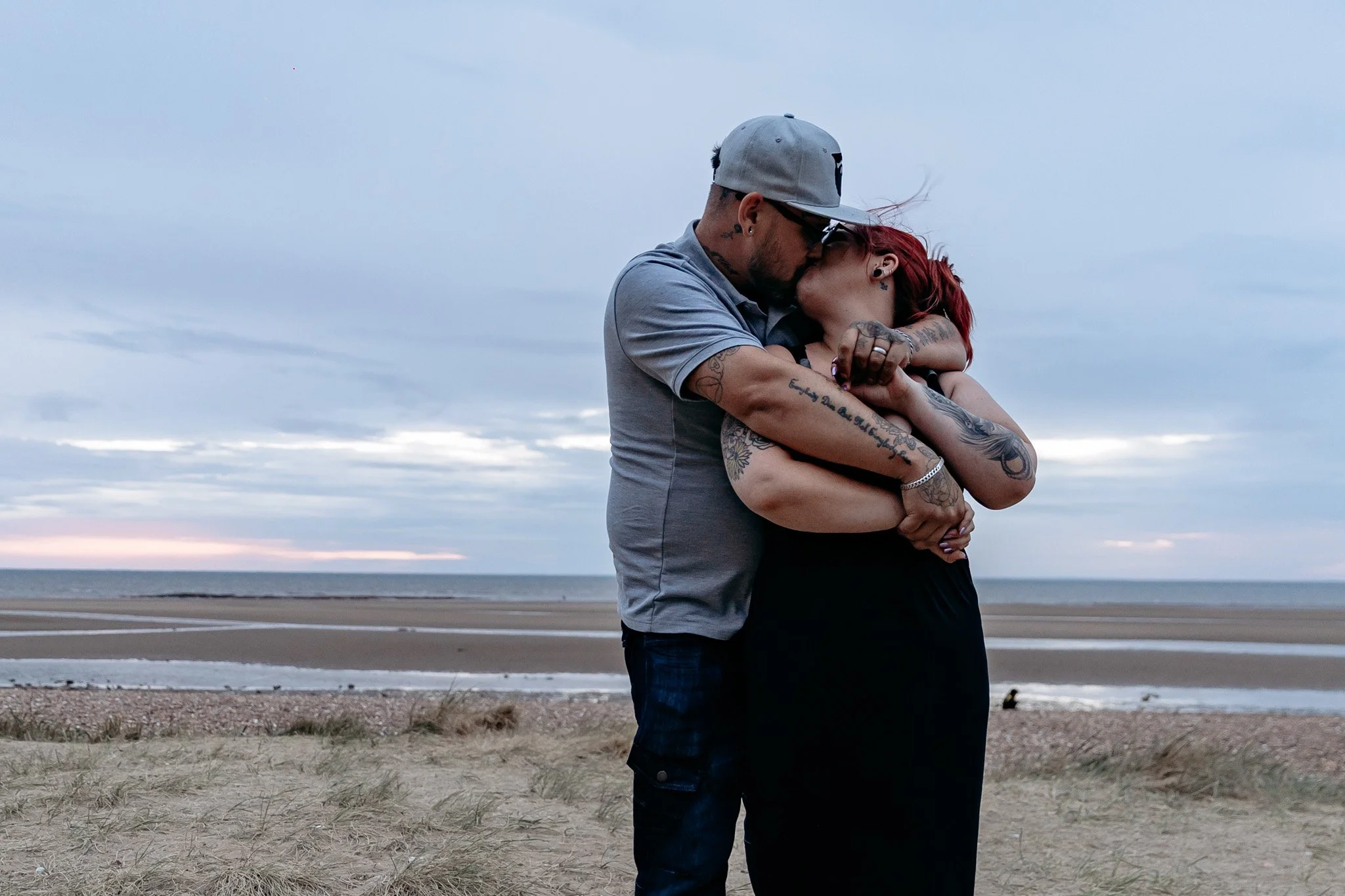 A couple kissing and embracing on a beach at sunset. The man is wearing a gray cap and gray shirt, and the woman has red hair and tattoos on her arms. The ocean and cloudy sky are in the background.