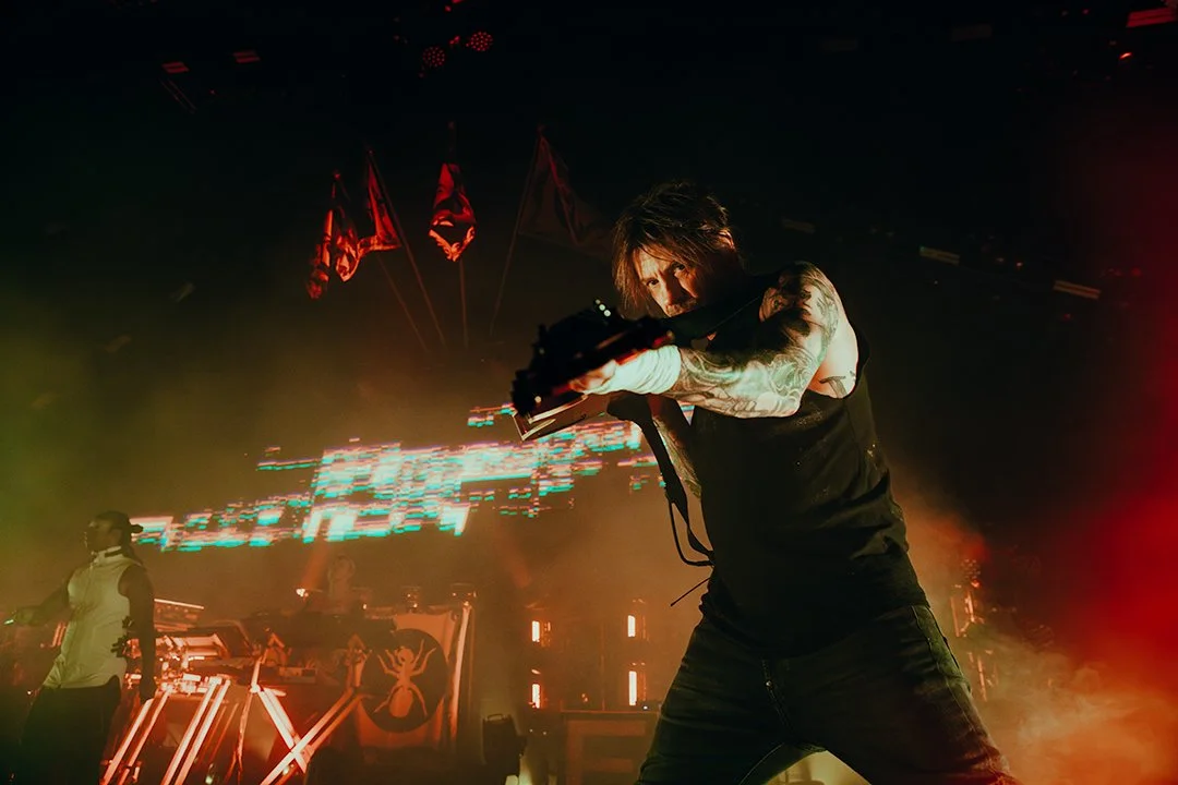 A musician with tattoos on his arms performing on stage, pointing a gun towards the camera, with red flags in the background and colorful lights.