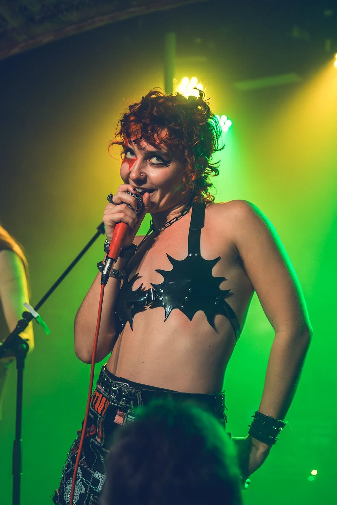 A female singer with curly red hair performing on stage, holding a microphone, wearing a black spiked bra, and black and red patterned pants with chains, under colorful stage lighting.