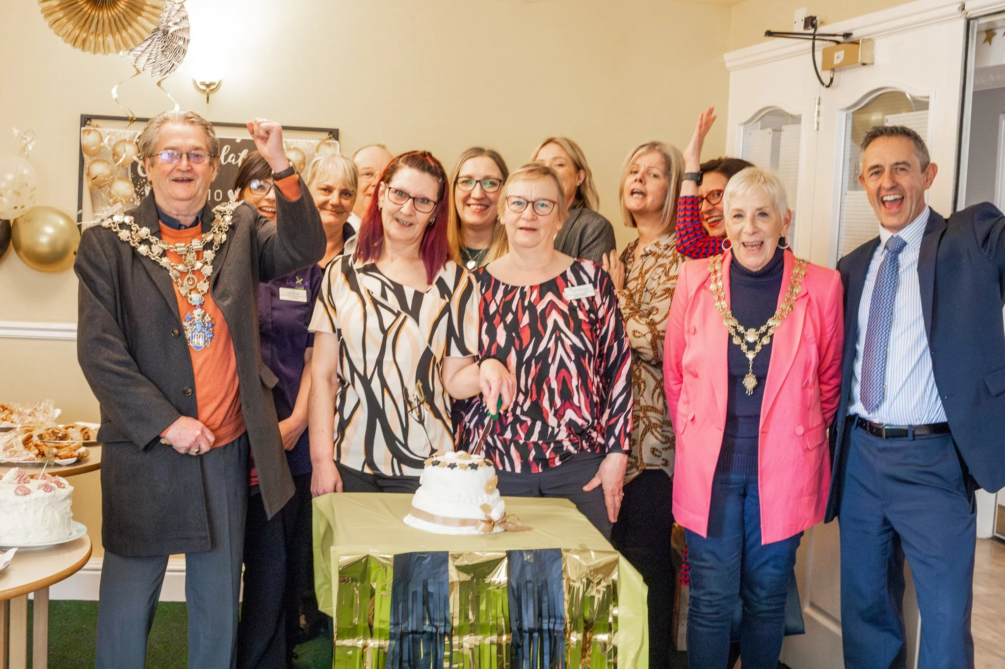 A group of people celebrating a birthday or special occasion, with a cake and balloons in a decorated room.