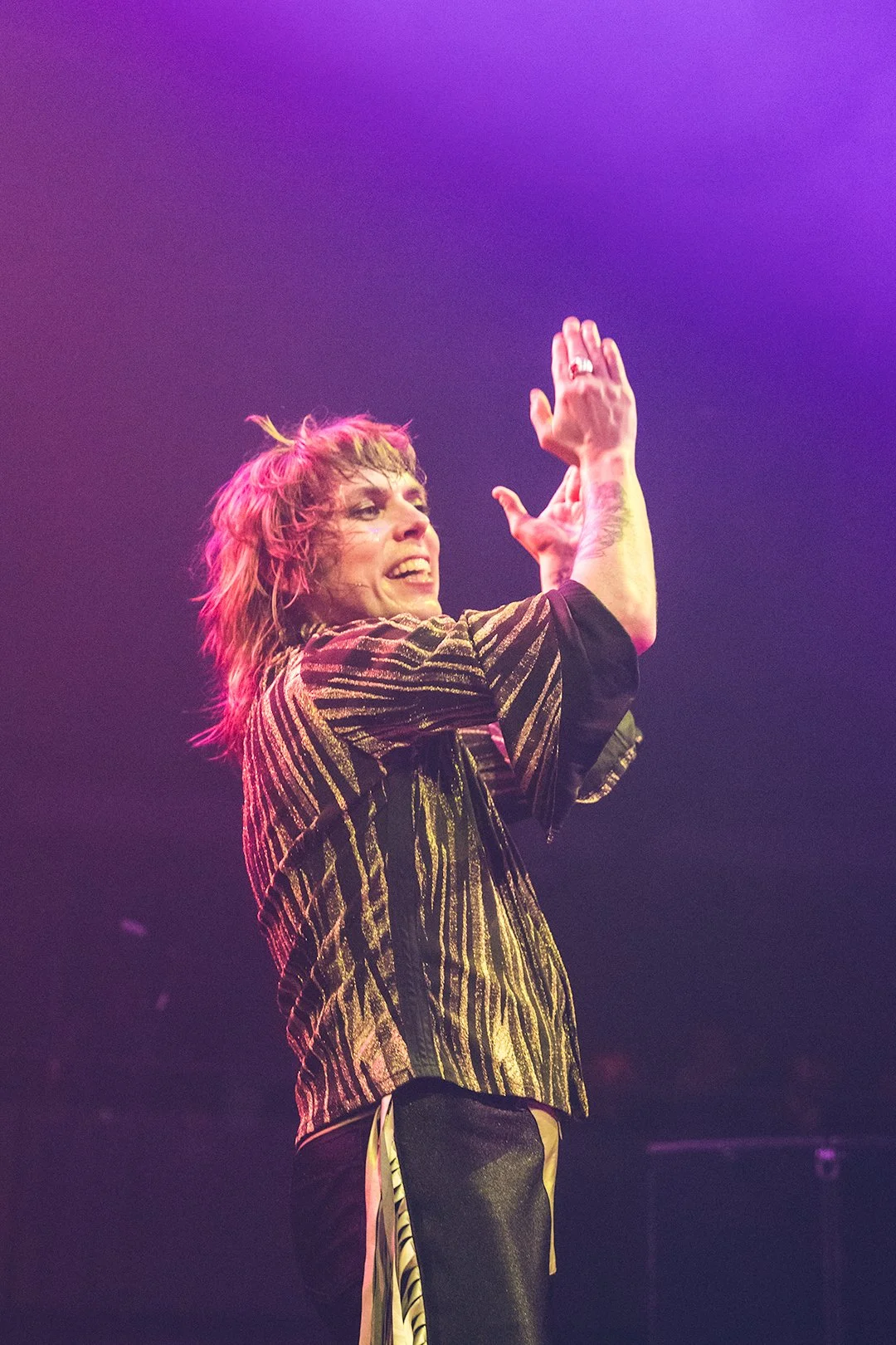 Performer on stage clapping, wearing a striped shiny jacket, with colorful stage lighting.