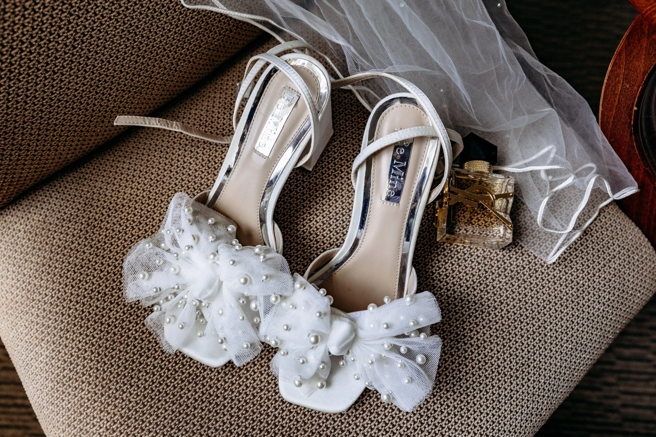 White high-heeled shoes decorated with large pearl-embellished bows, placed on a textured brown fabric surface, alongside a small perfume bottle and a white veil or fabric.