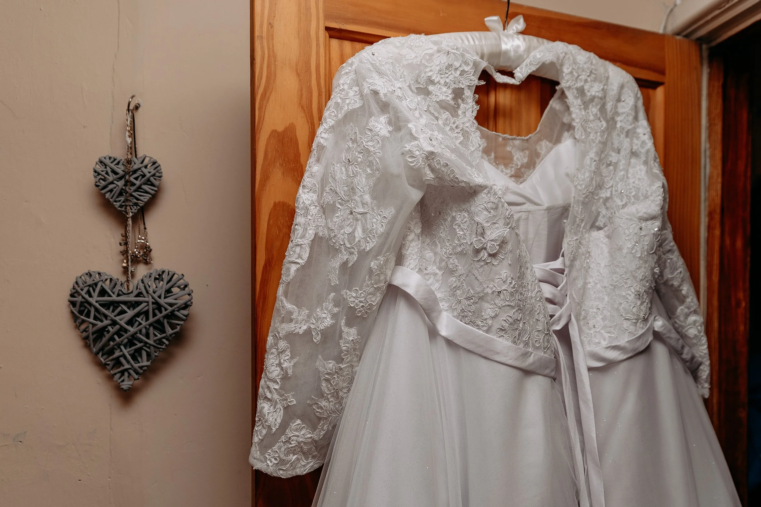 Two white wedding dresses hanging on a wooden door, with lace and satin details, and a hanging black and white heart-shaped decoration on the wall.