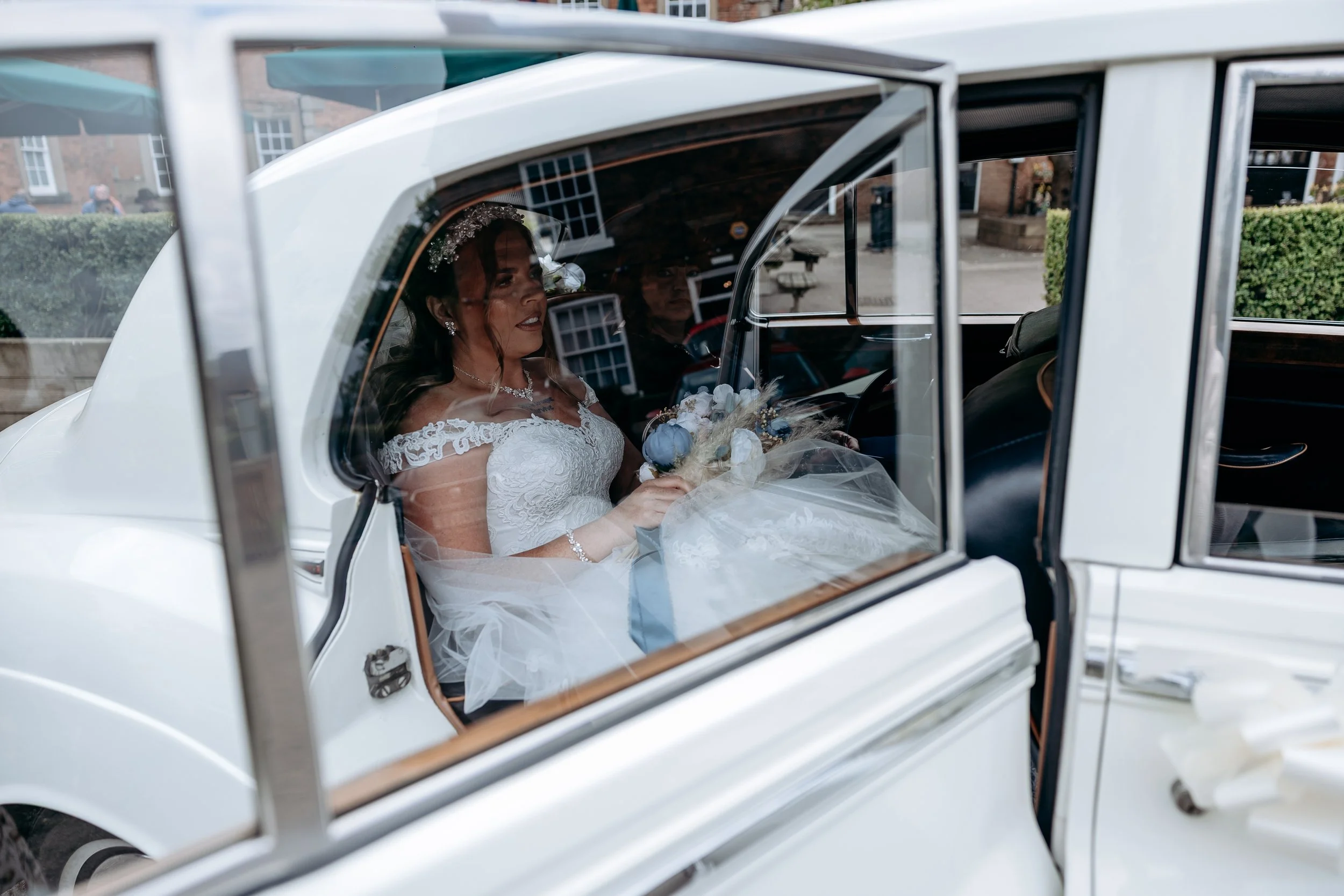 A bride sitting inside a vintage white car holding a bouquet, seen through the car window.