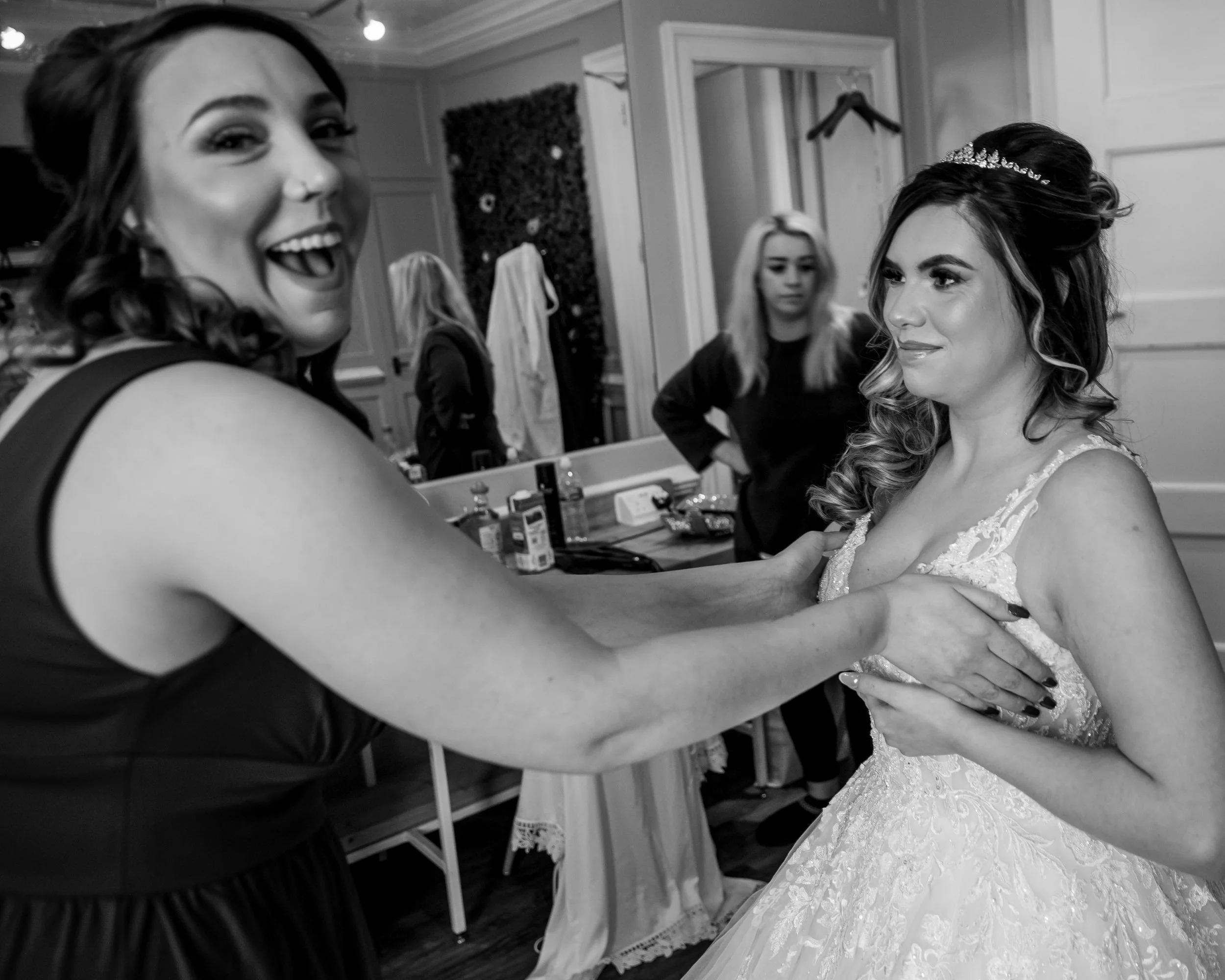 A woman in a dark dress laughs and holds hands with a bride in a lace wedding gown in a dressing room, while two other women are visible in the background.