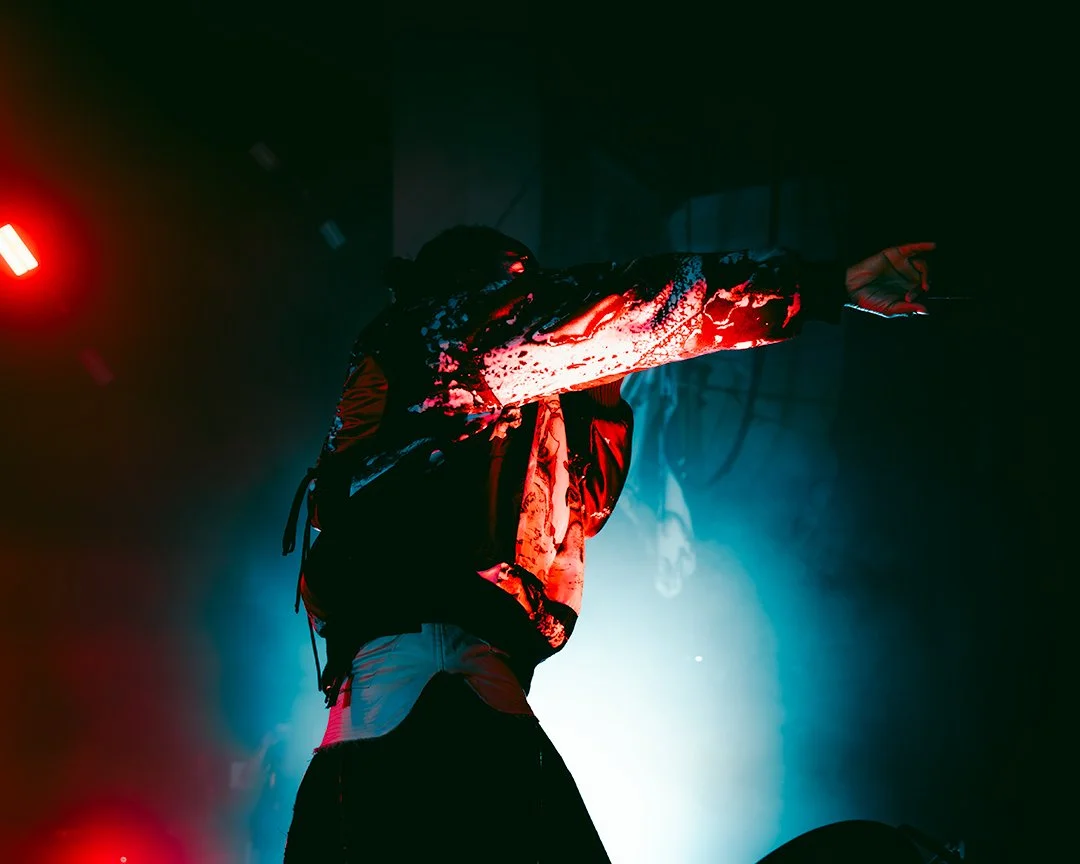 A performer on stage pointing forward, illuminated by red and blue stage lights, wearing a patterned jacket and a backpack.