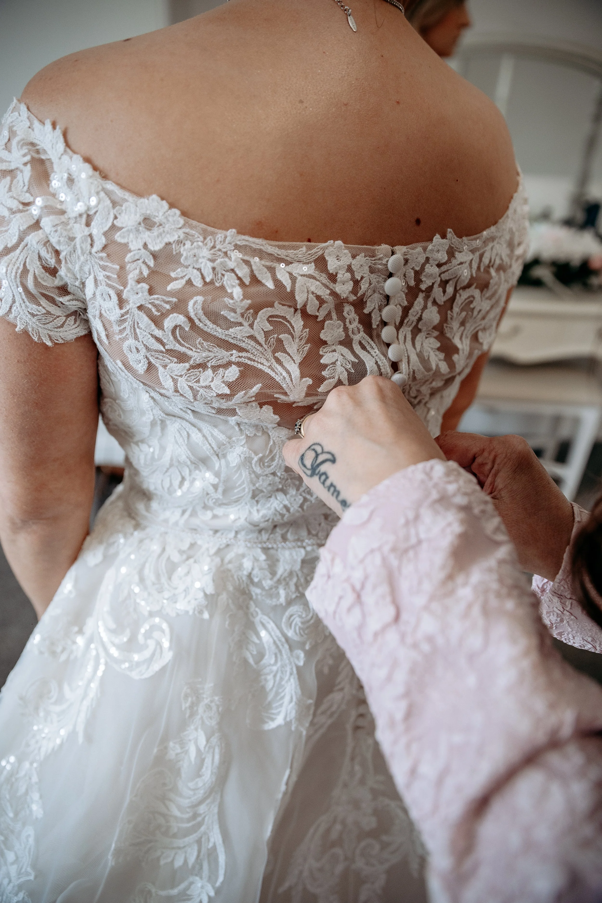 A bride in a white lace wedding dress is being buttoned up at the back by another person with a tattoo on their hand.