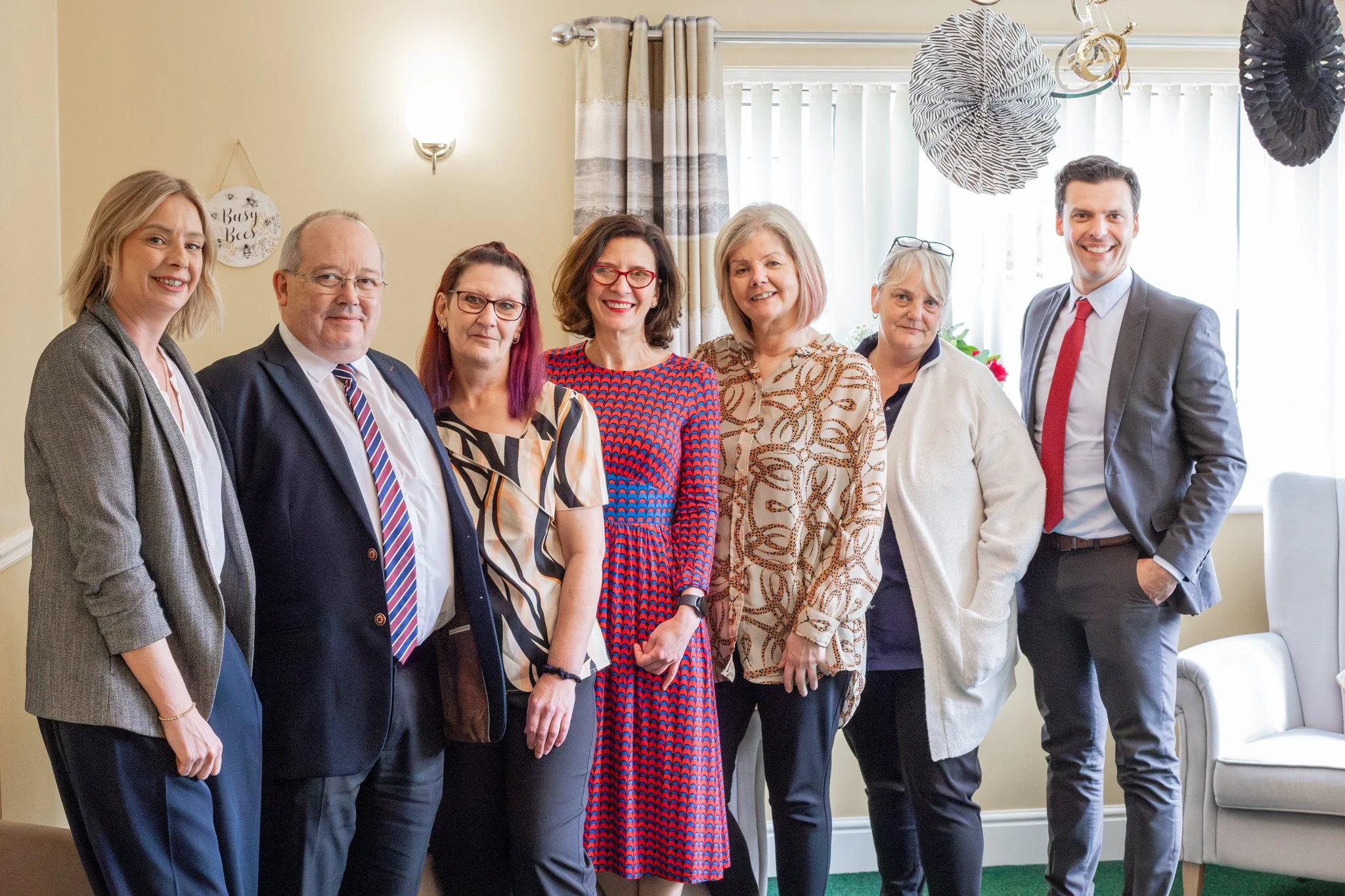 A group of eight people, six women and two men, standing together indoors for a group photo. They are dressed in business casual and formal attire, smiling, in front of decorated curtains and balloons.