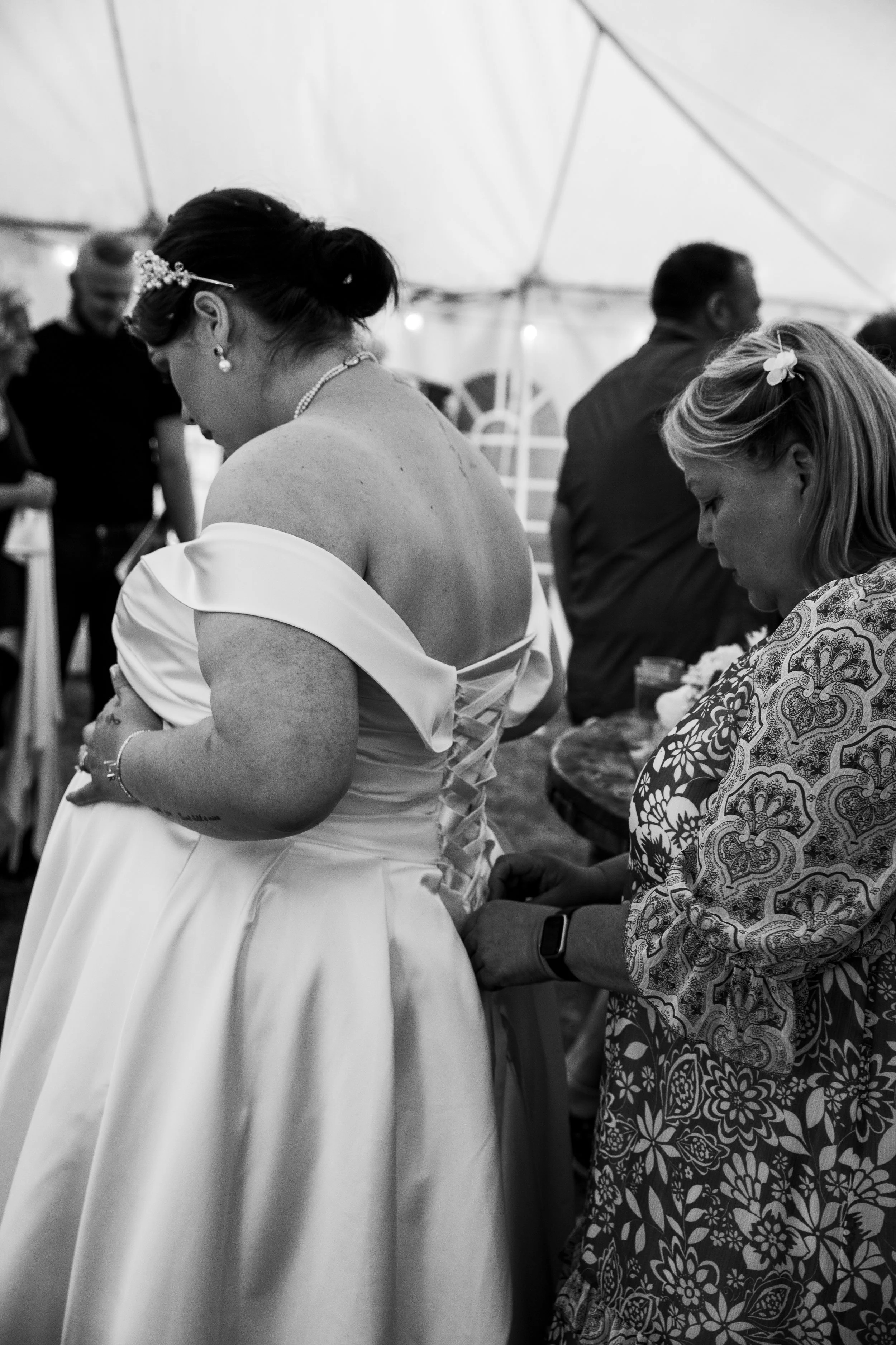 A bride in a wedding dress is being helped to tie her gown's lace-up back by a woman in a floral dress at a wedding reception.