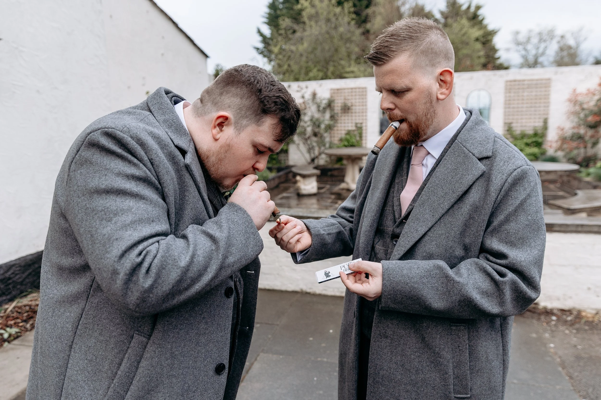 Two men in gray coats smoking cigars outdoors, one leaning forward and the other holding a small box.