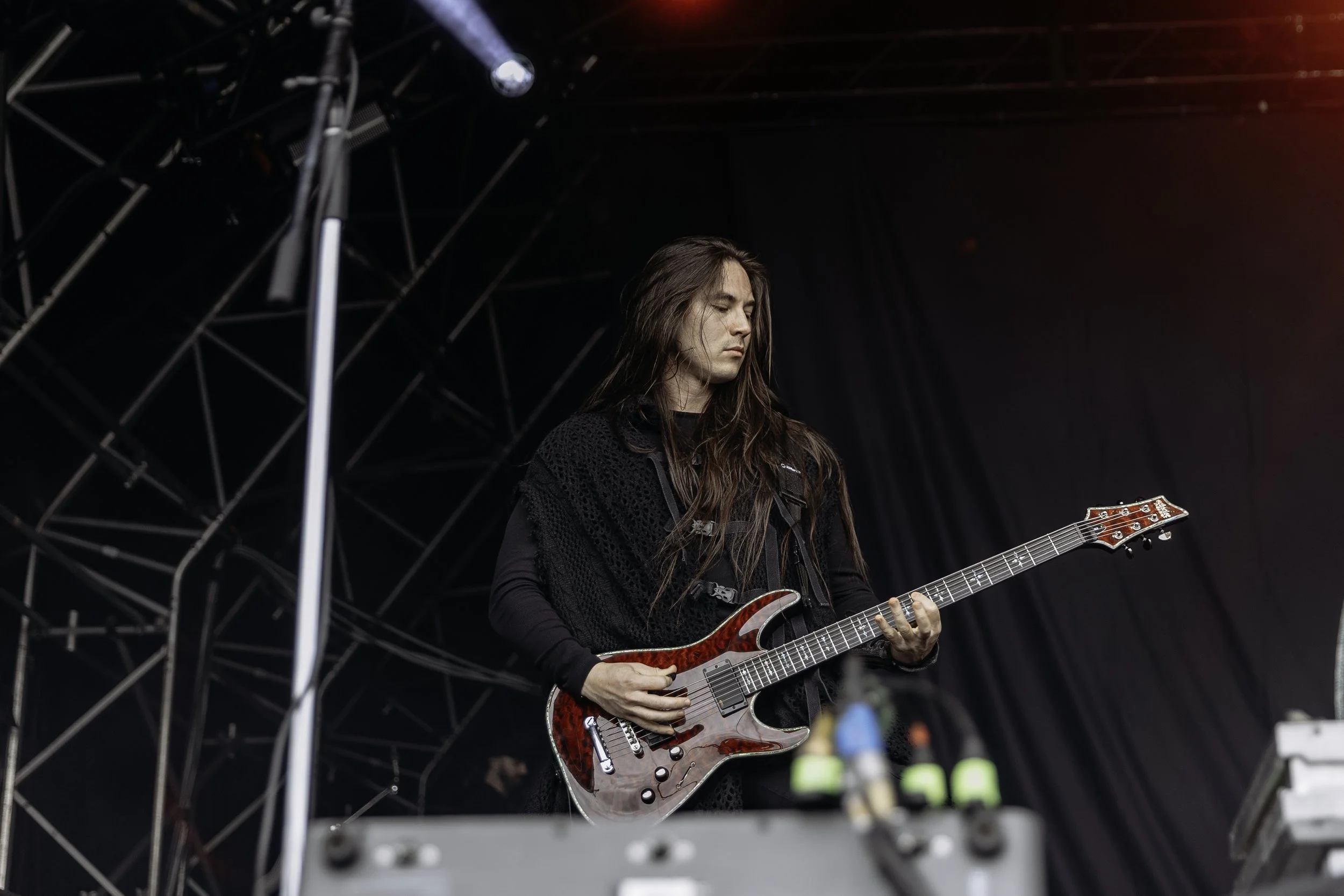 A long-haired musician playing a red electric guitar on stage with dark background and stage equipment.