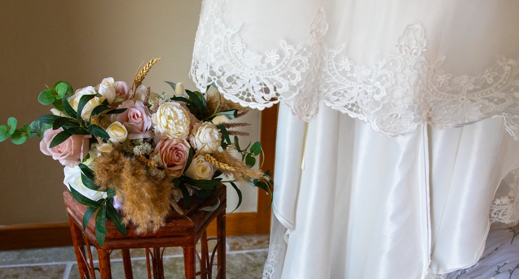 A bouquet of pink and white roses with greenery and dried brown accents on a wooden stand beside a white lace and silk wedding dress.