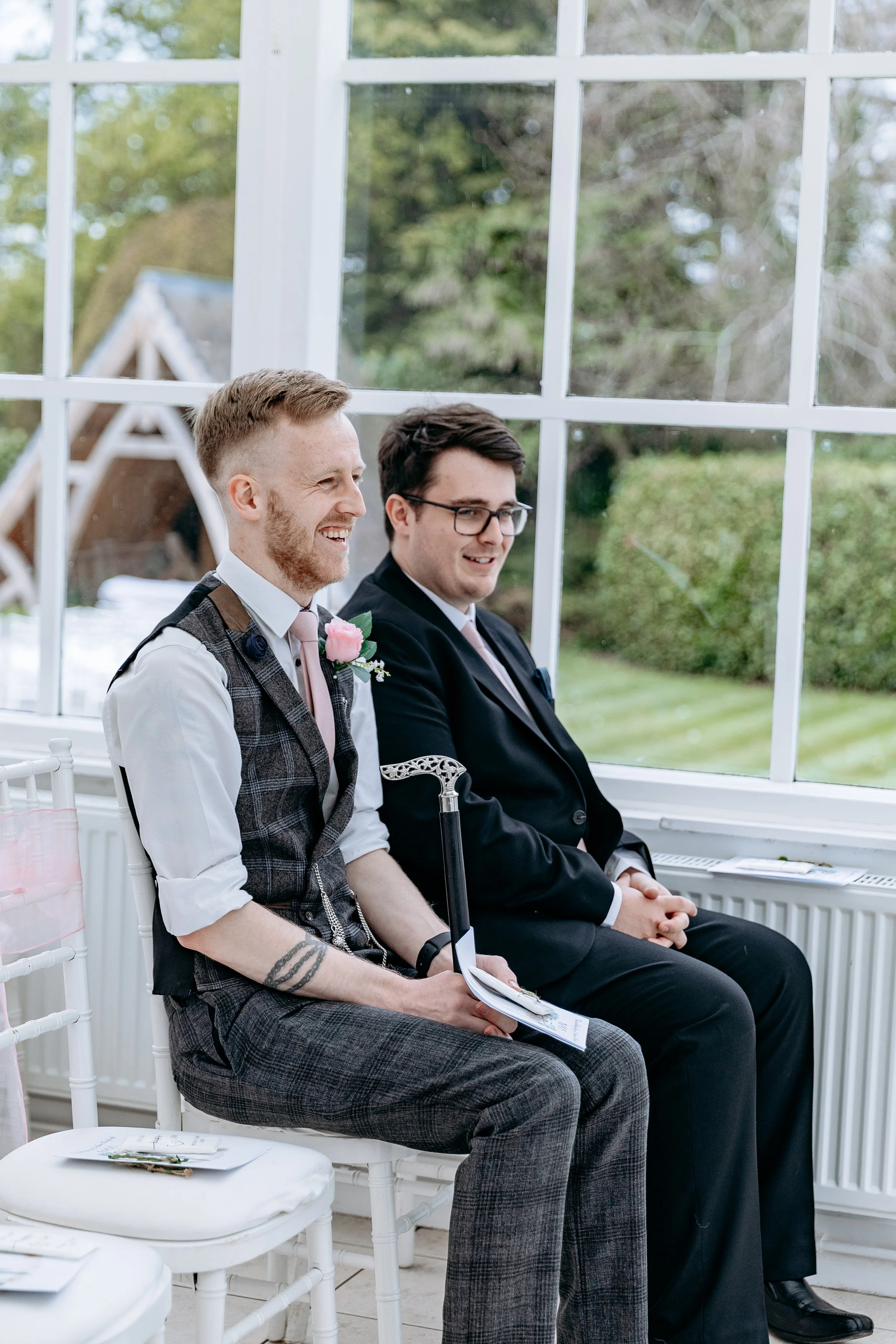 Two men dressed in formal attire sitting inside by a large window, smiling during a wedding ceremony.