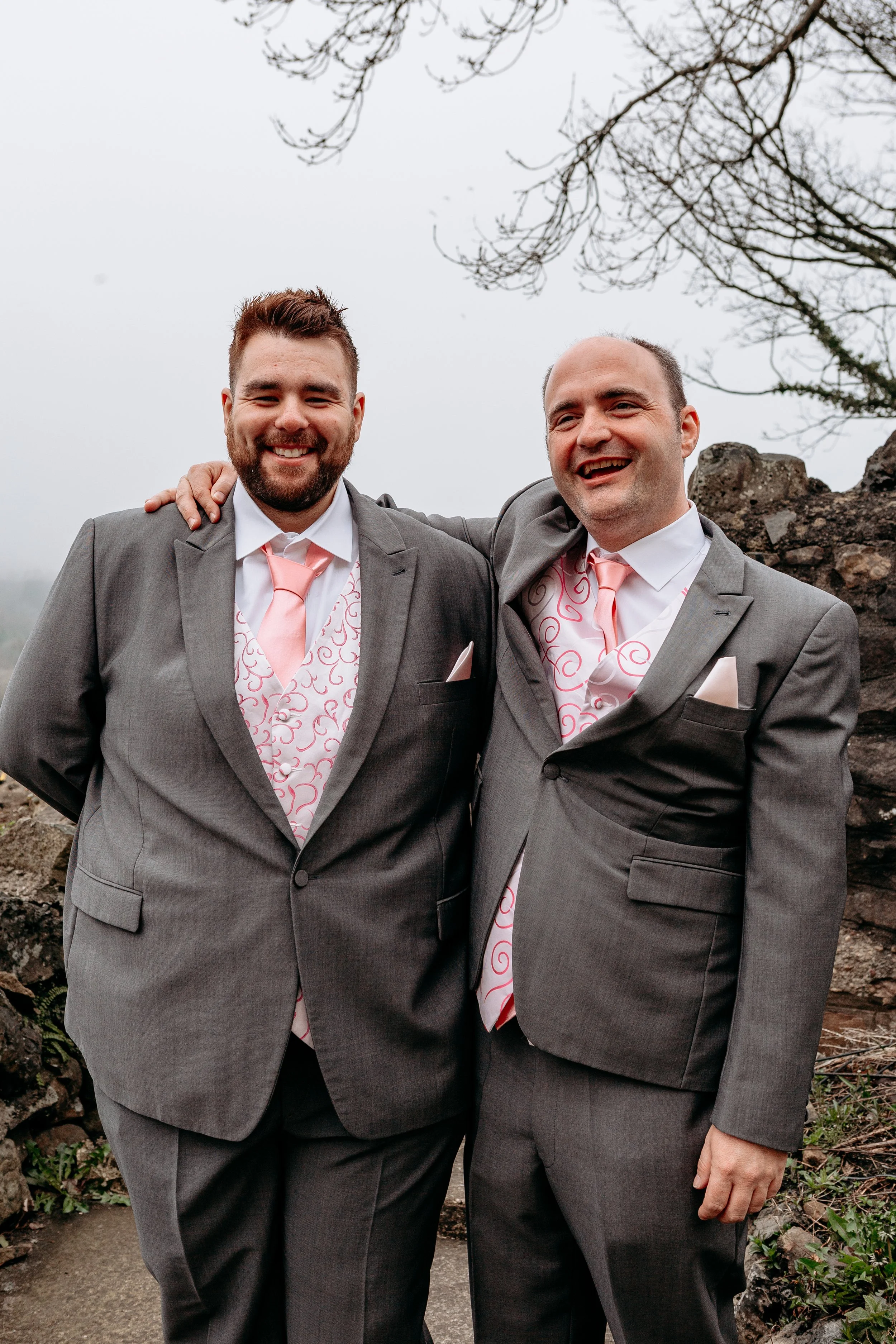 Two men in gray suits with pink ties and vests, smiling, standing outdoors with their arms around each other.