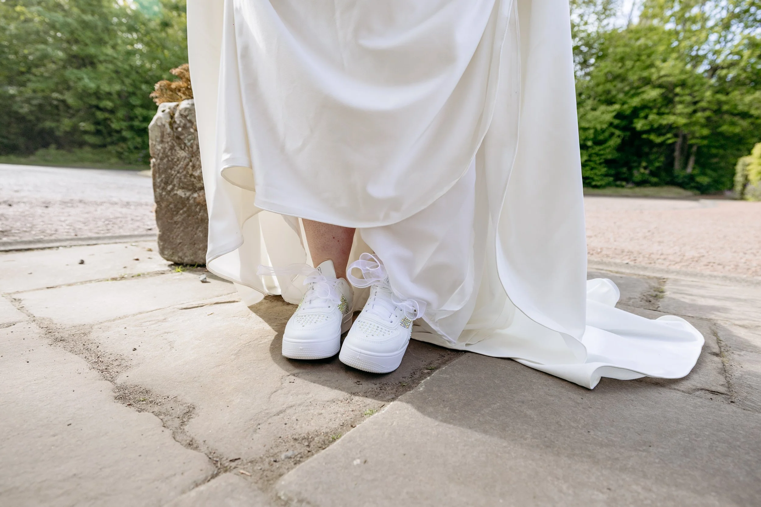 Close-up of a person in a wedding dress wearing white sneakers with decorative elements, standing outdoors on stone pavement.