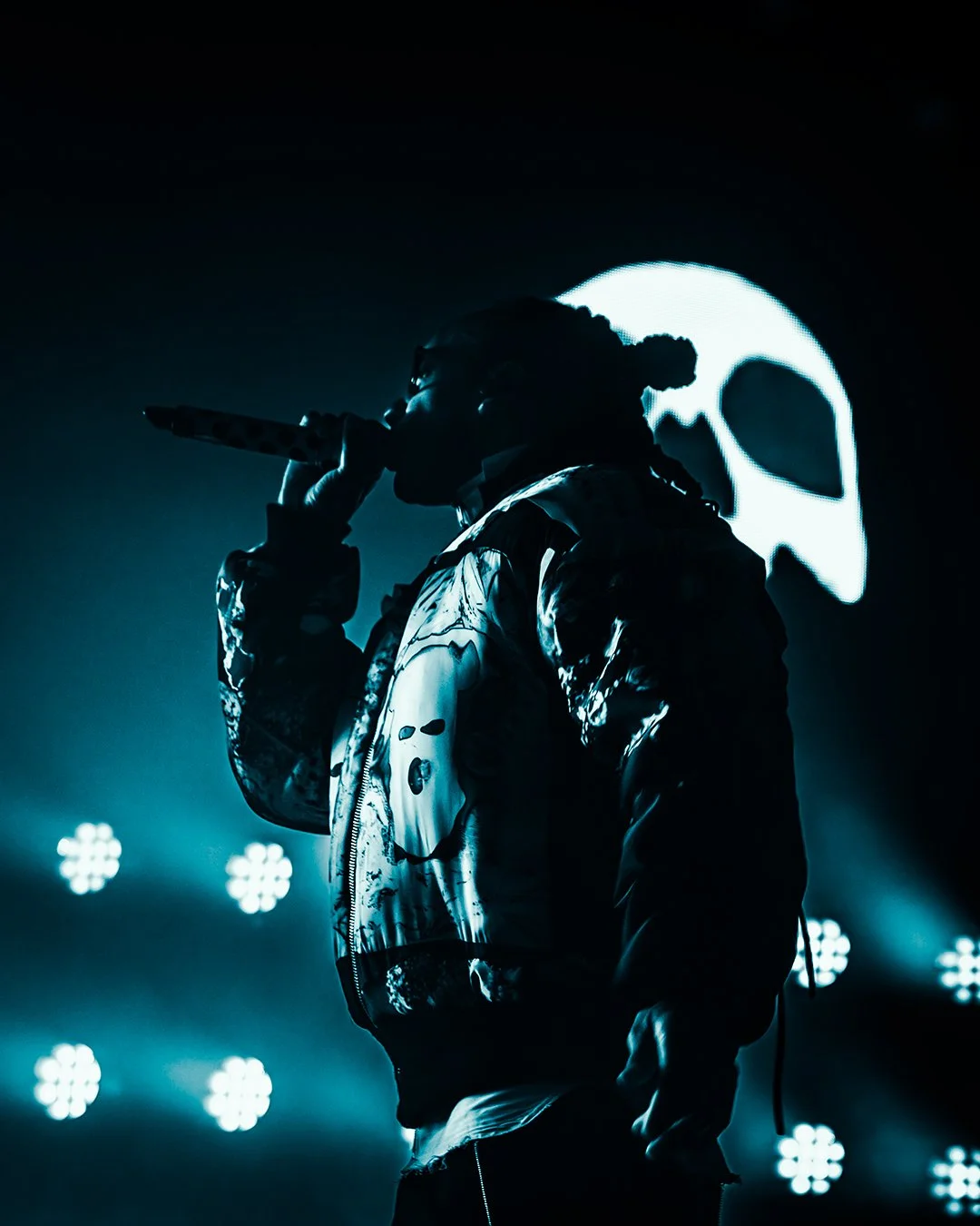 A silhouette of a performer on stage holding a microphone, with a large illuminated skull behind them and multiple bright lights in the background.