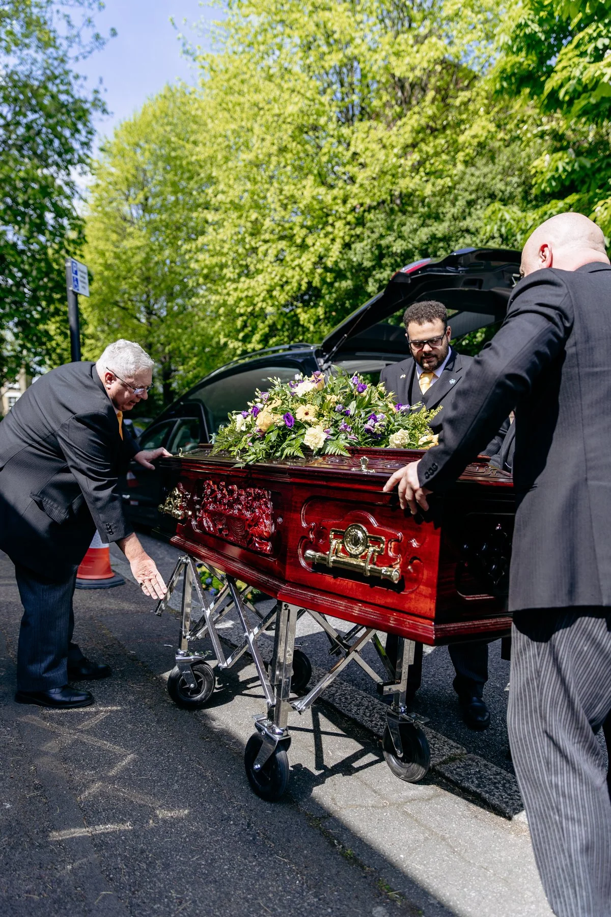 Three men in suits placing a polished wooden casket with a floral arrangement on top into a hearse outside on a street with green trees in the background.
