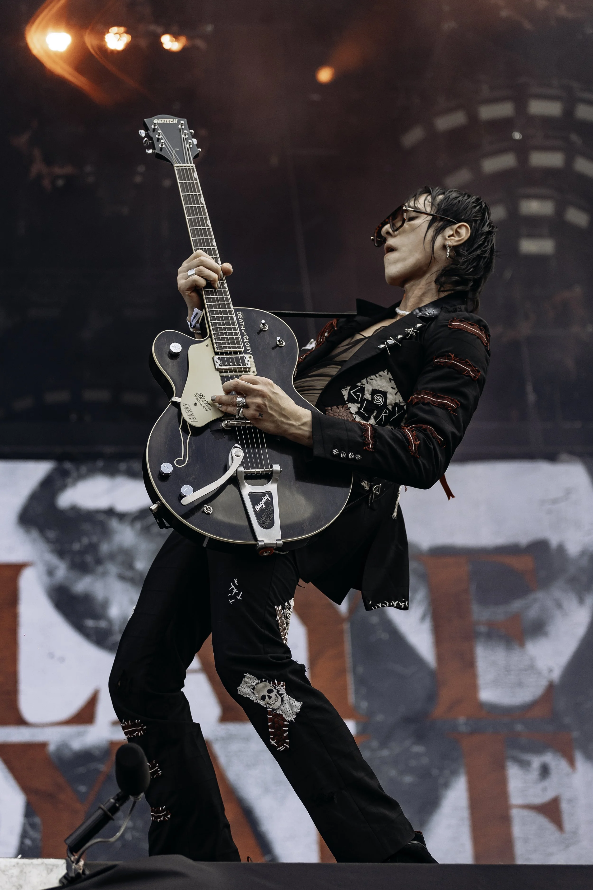 A male guitarist performing on stage with black hair, sunglasses, and Gothic-style clothing, playing a black electric guitar with white accents, during a concert.