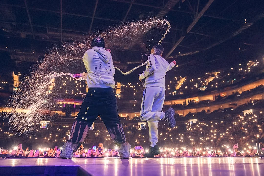 Two performers on stage spraying water with a crowd of fans holding phones in an arena setting.