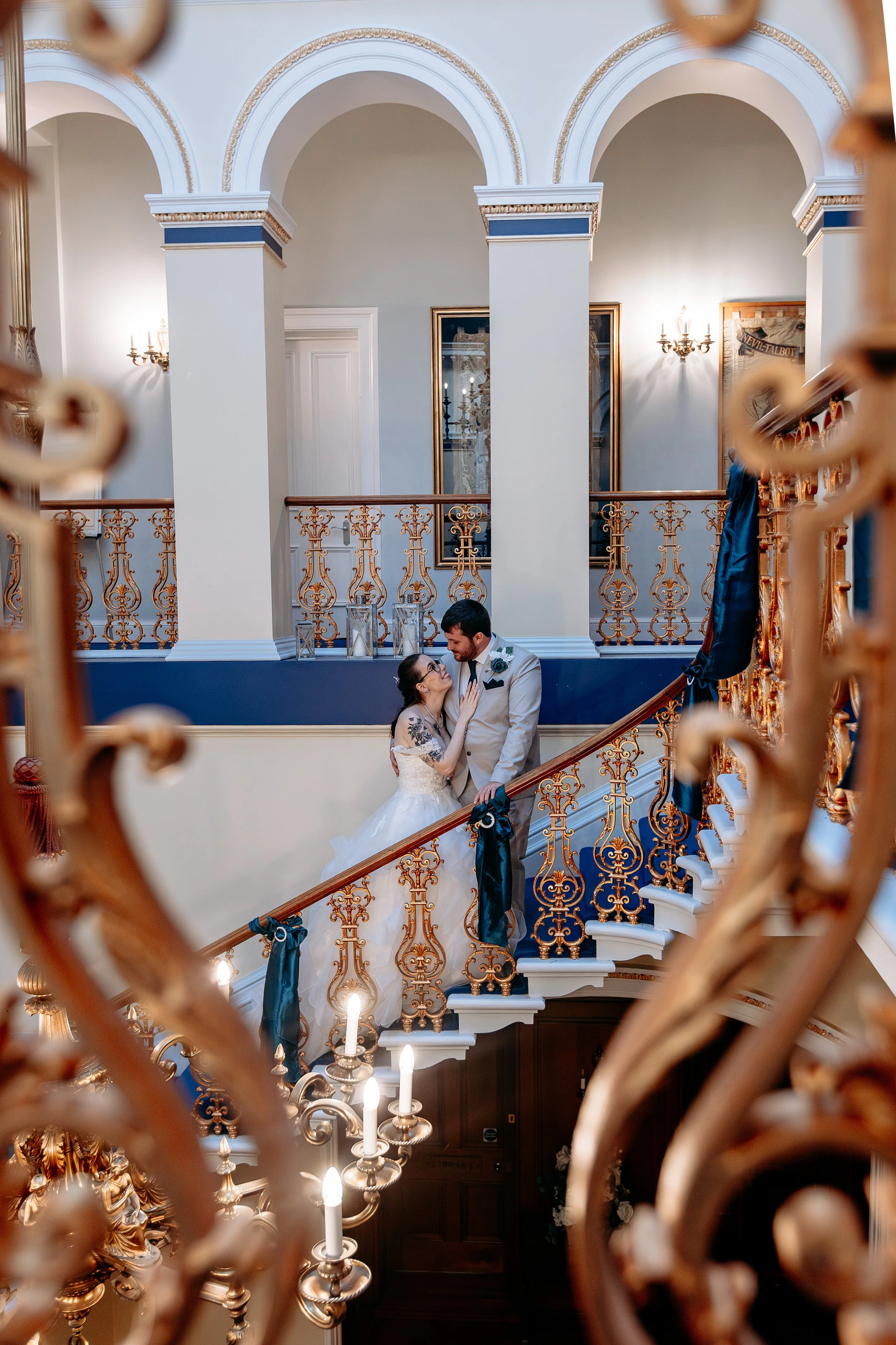 A bride and groom standing on a grand staircase, intimately gazing at each other, with ornate gold railings and elegant decor in the background.