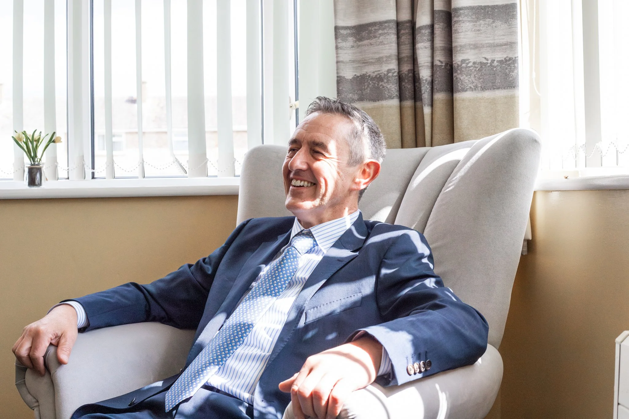 A middle-aged man in a business suit sitting in a beige armchair, smiling and relaxed in a bright room with large windows and curtains.