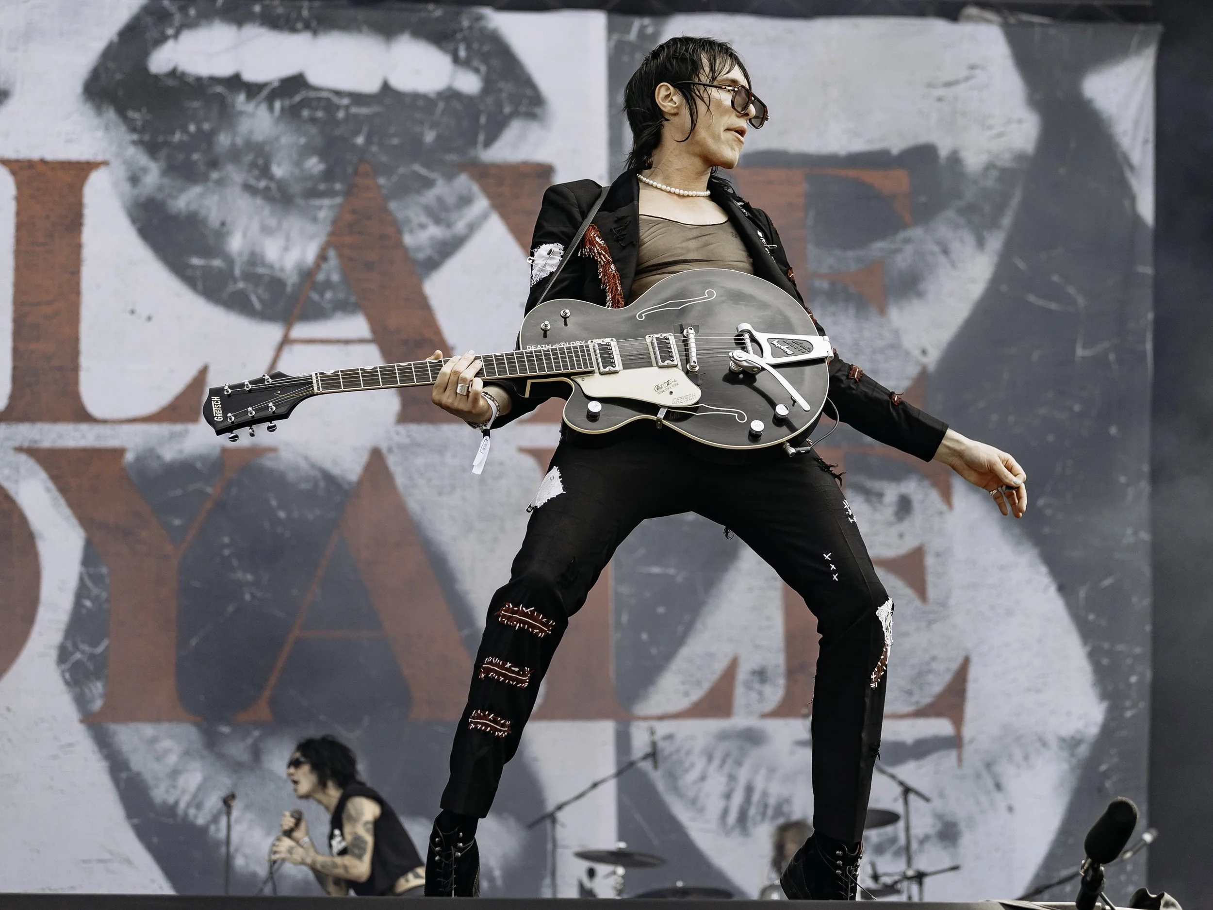 A woman playing an electric guitar on stage with a large backdrop featuring a black-and-white face and red text. Another singer is seen in the background.