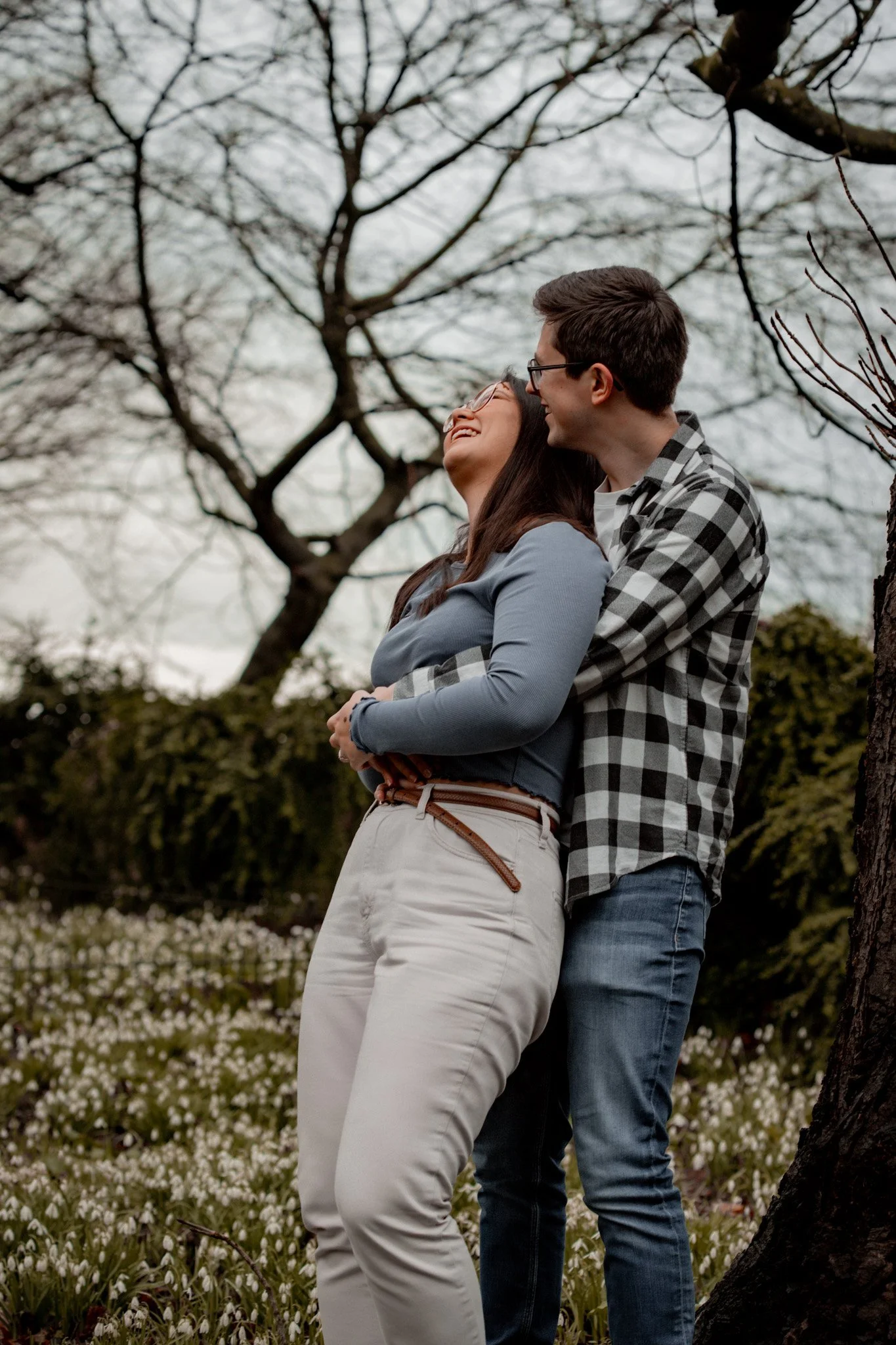 A young couple smiling and hugging outdoors with trees and white flowers in the background.