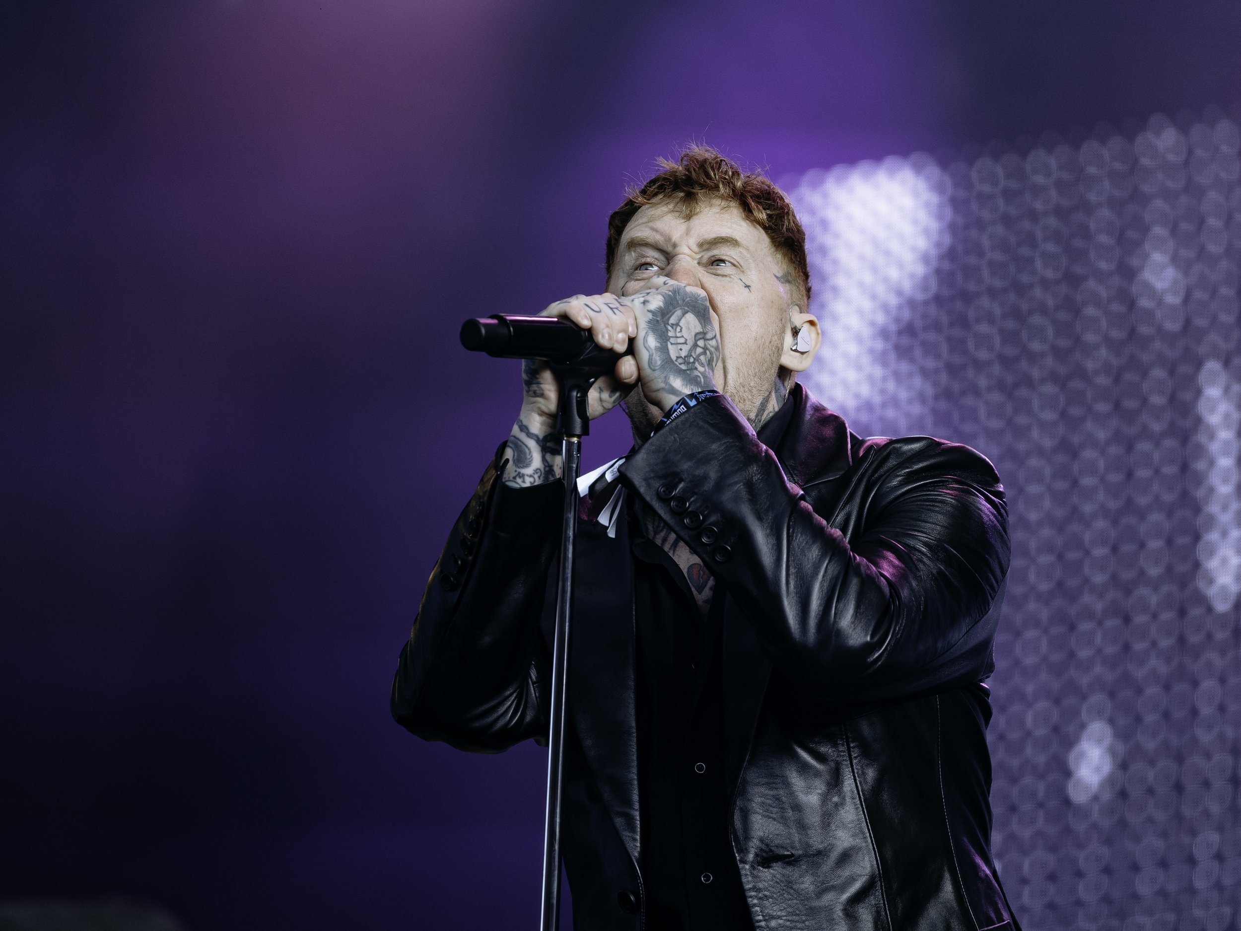 A male singer with tattoos and a leather jacket passionately singing into a microphone on stage, with vibrant purple and blue lights in the background.