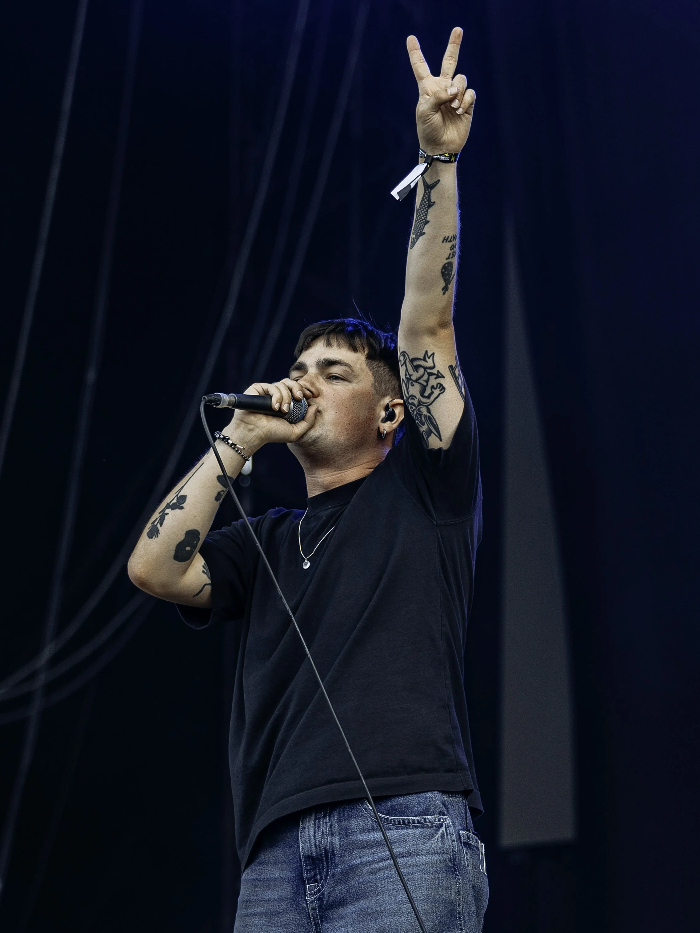 A male singer with tattoos performing on stage, holding a microphone in his right hand, with his left hand raised making a peace sign.