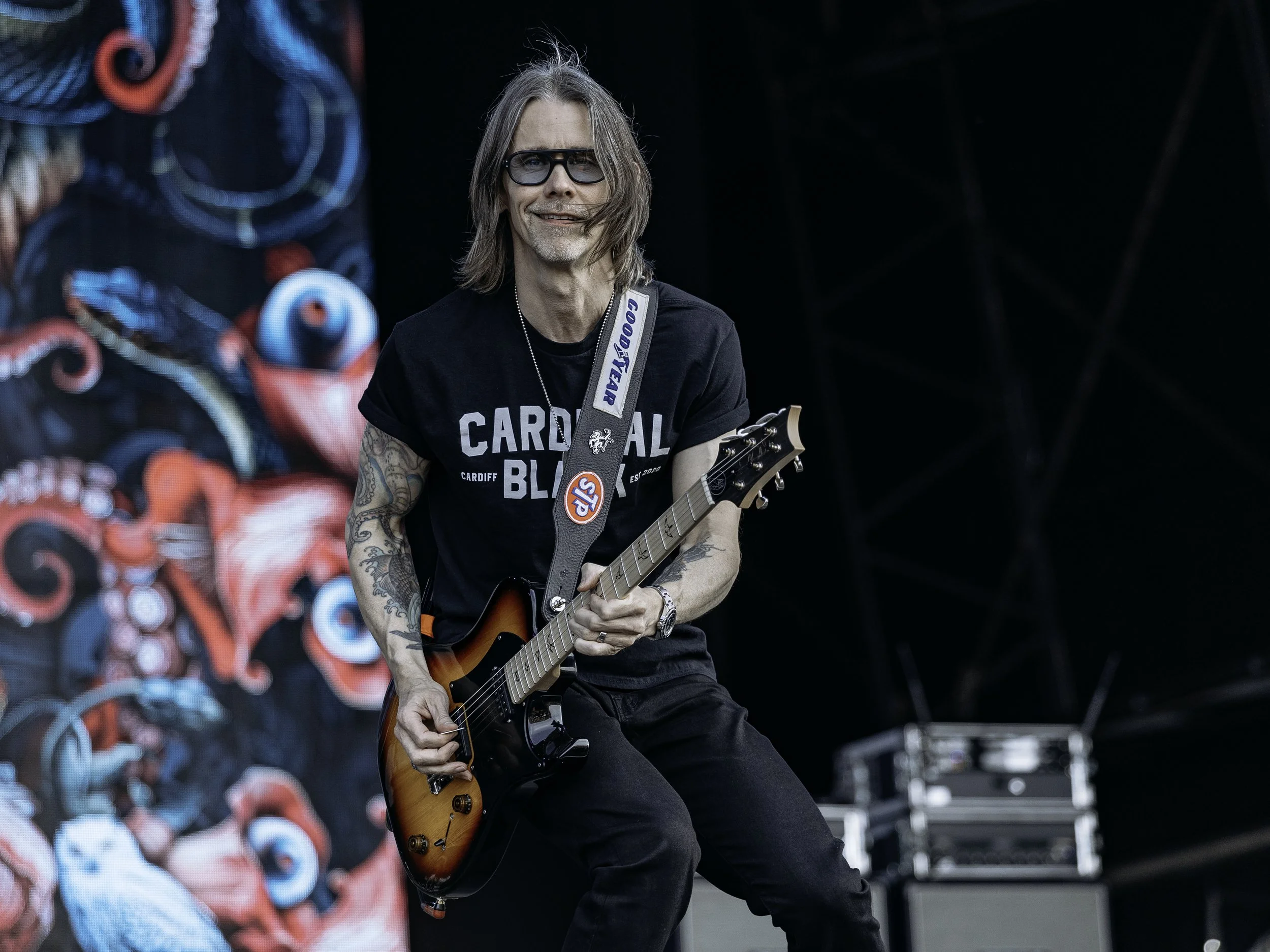 A male musician with shoulder-length hair, glasses, and tattoos, wearing a black "Cardiff Blues" T-shirt, playing an electric guitar on stage with a colorful backdrop and sound equipment in the background.