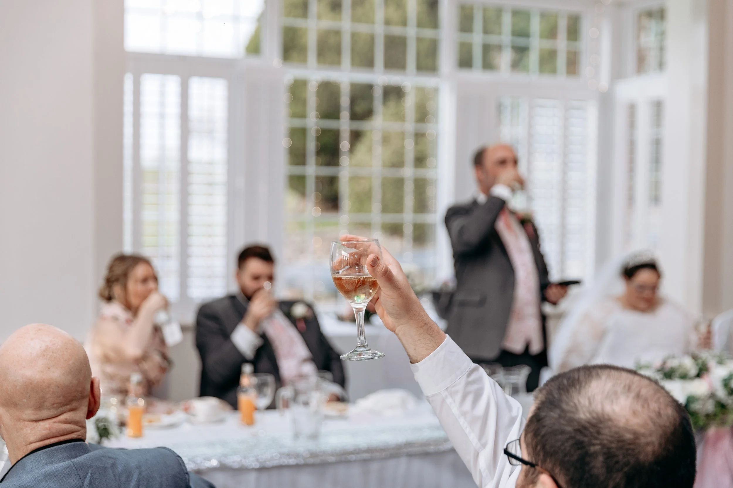 Person holding a glass of pink wine at a wedding reception, with seated guests and a speaker in the background.