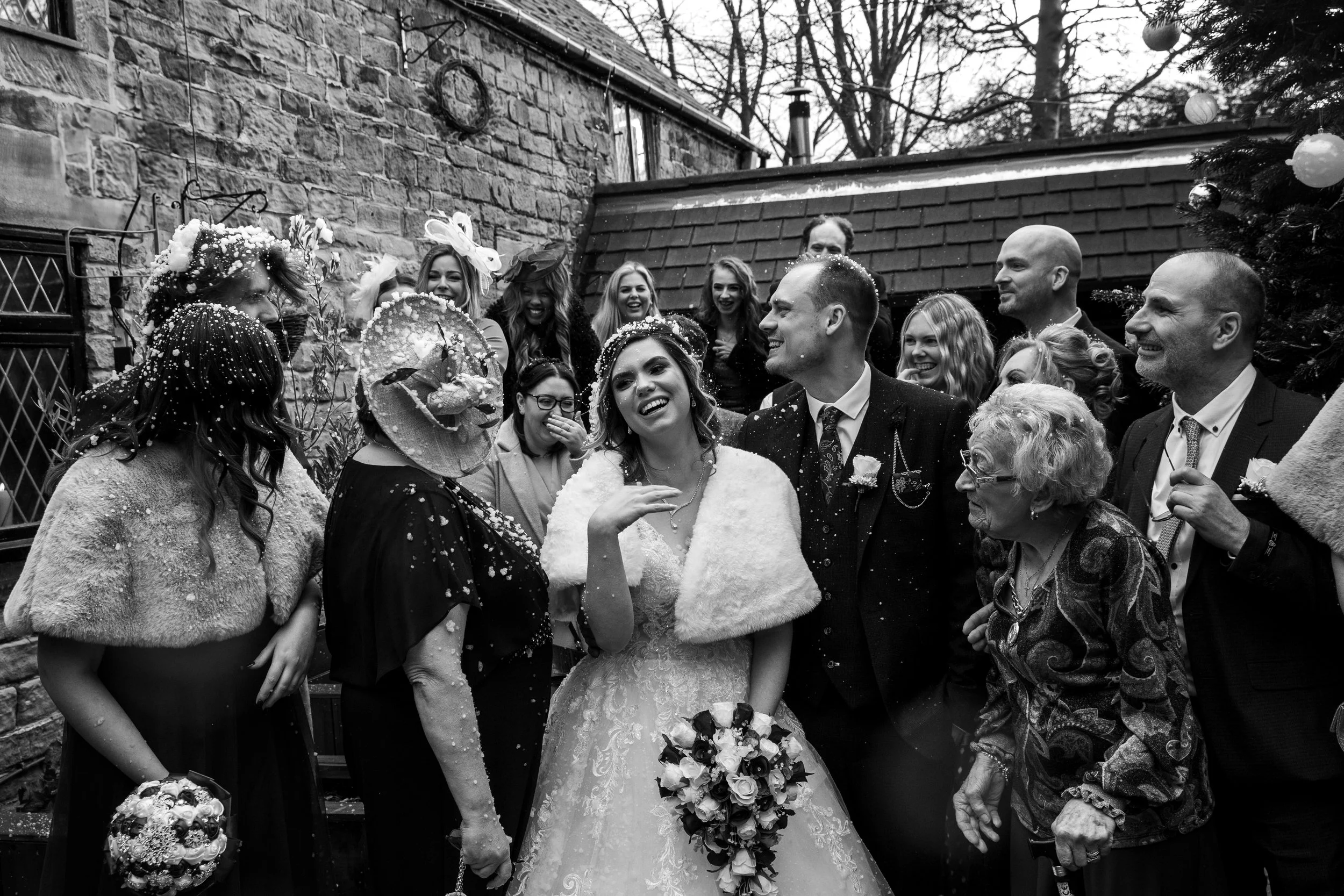 Black and white image of a bride and groom surrounded by smiling wedding guests, celebrating outside near a brick building and a decorated Christmas tree.