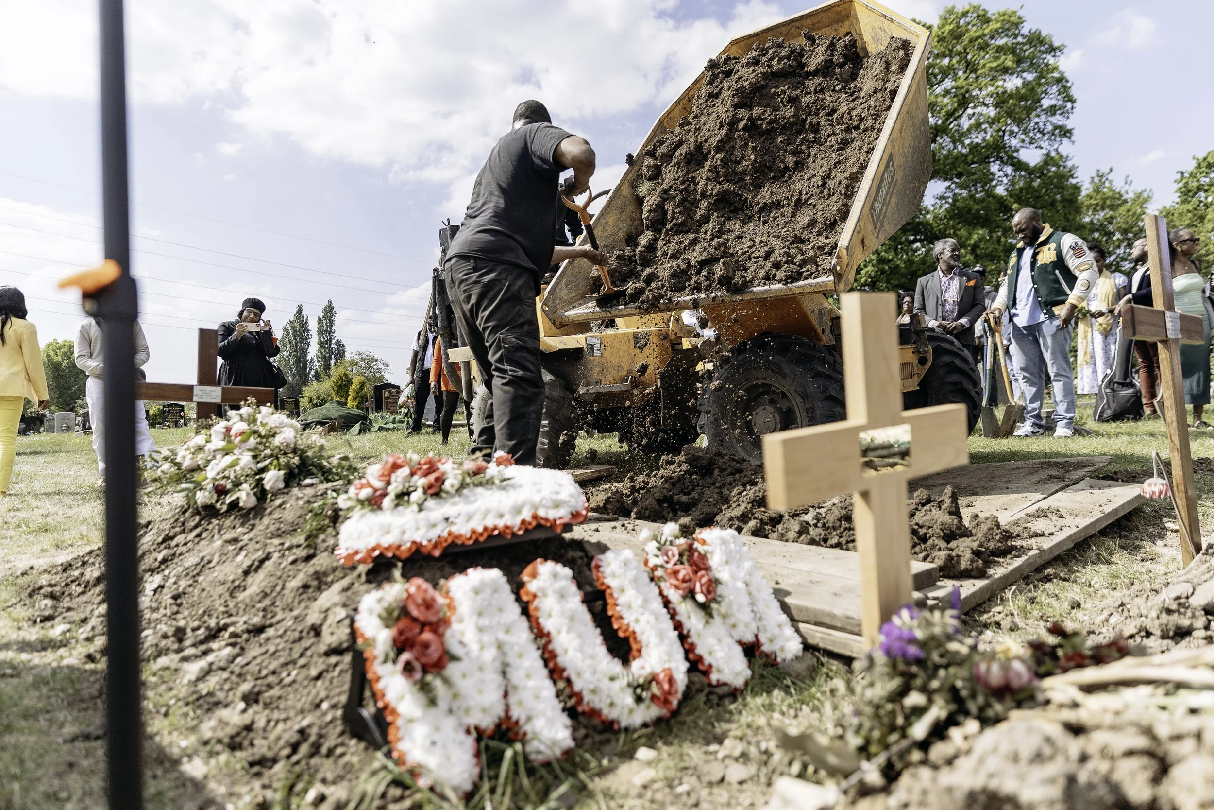 People participating in a gravesite funeral, with a man shoveling dirt into a grave and floral arrangements placed on top and nearby.