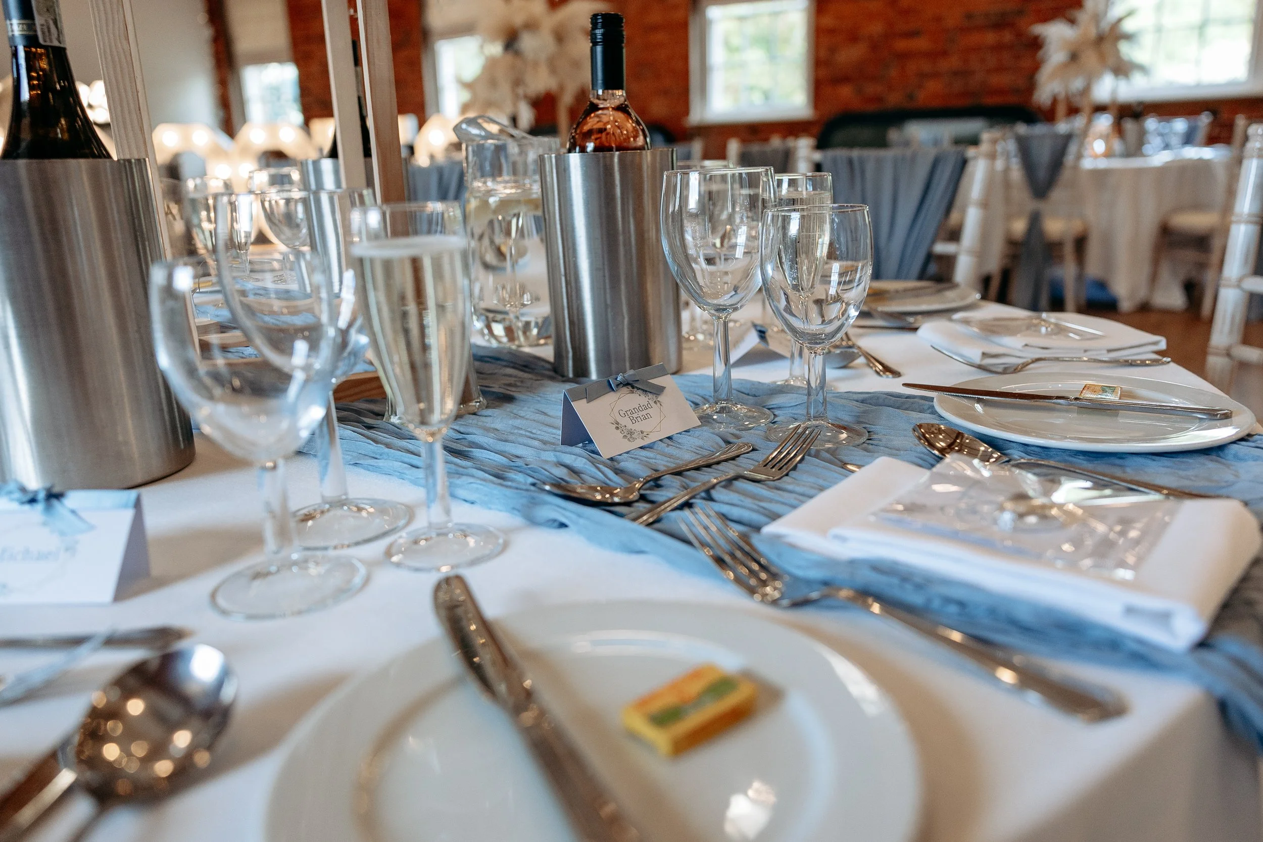 Elegant banquet table set with white plates, silverware, wine glasses, champagne flutes, and a blue table runner, with a small place card reading 'Grandad Brian' and a bottle of rosé wine in a silver wine holder.